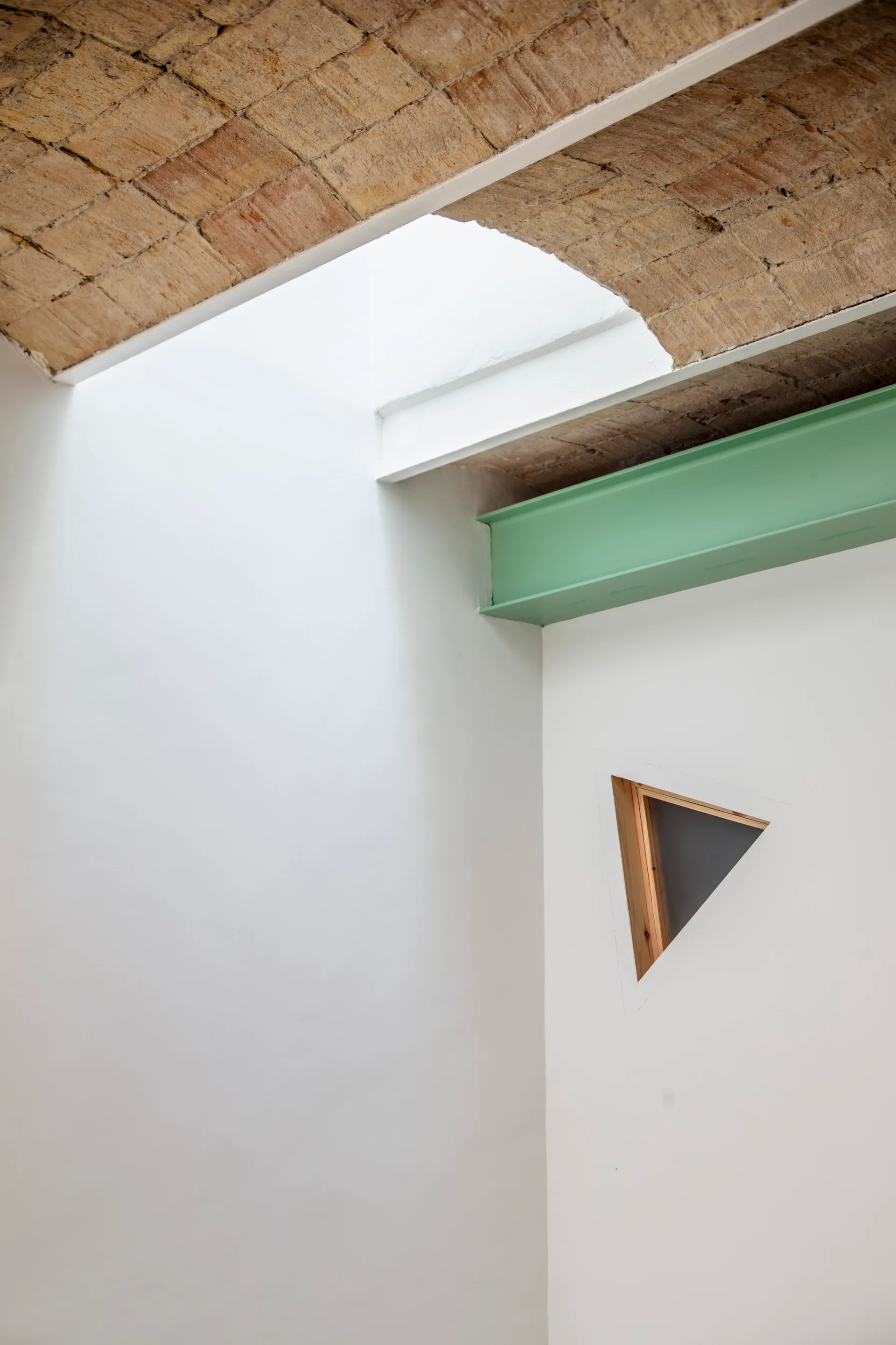 A white plaster wall features a small triangular wood-framed window beneath a green painted steel beam and an exposed brick vaulted ceiling.