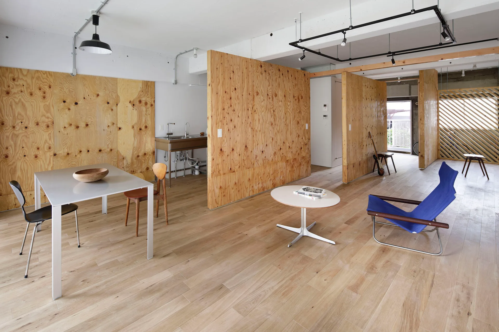An open-concept living area features raw plywood partition walls, light wood flooring, a stainless steel sink unit, and a blue canvas lounge chair.