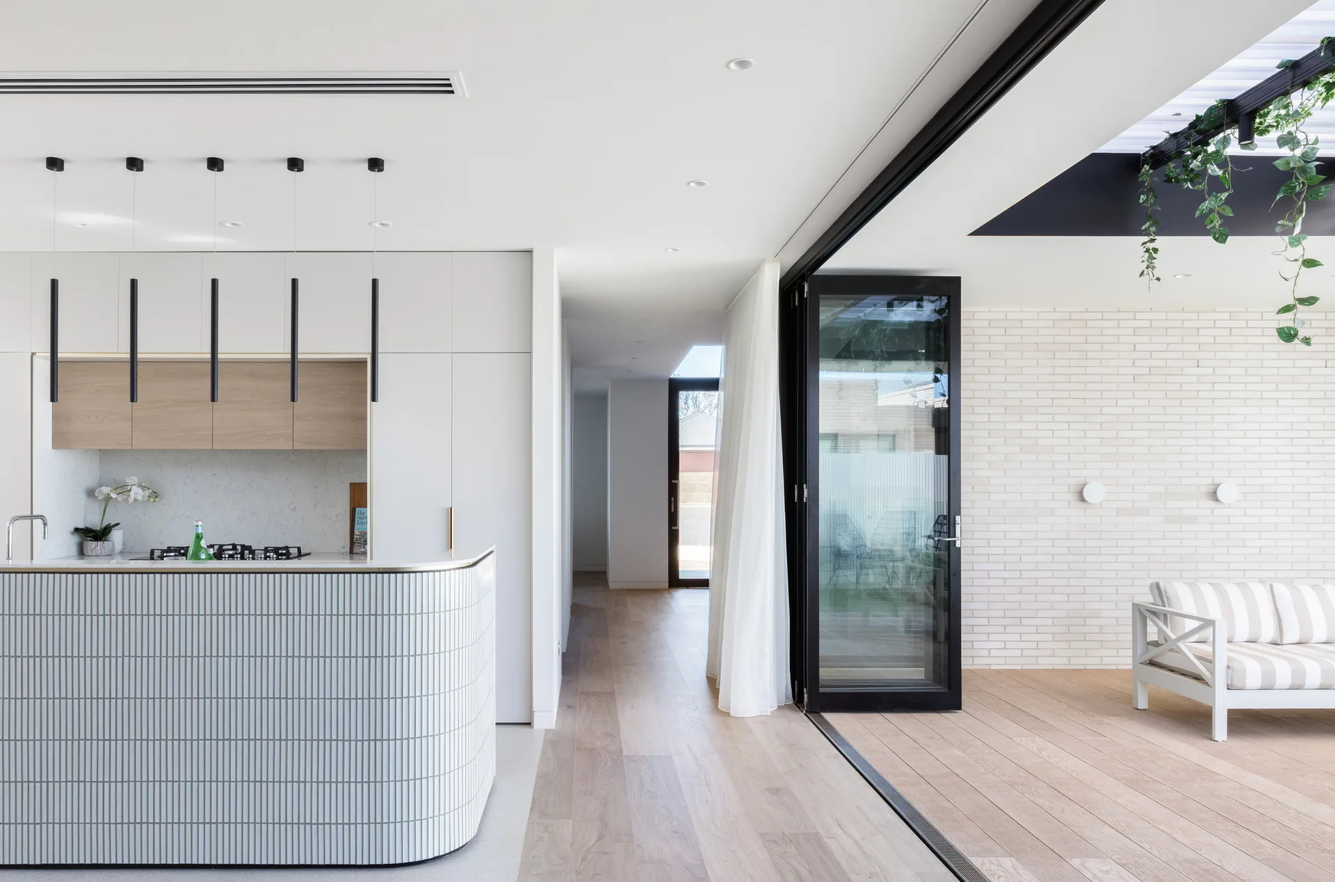 A modern white kitchen with a curved fluted island opens up to an outdoor patio with wood decking and a striped sofa.