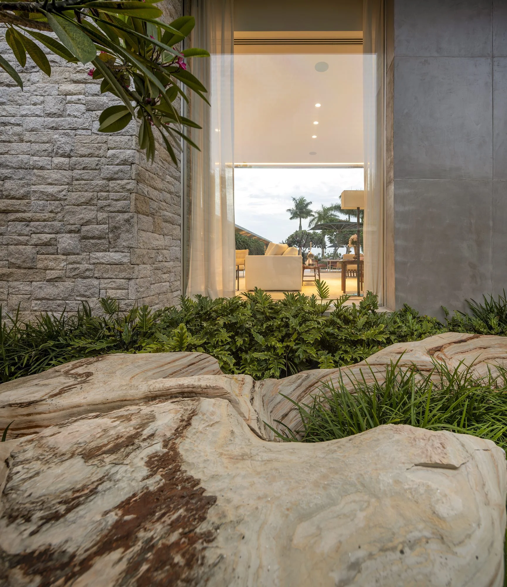 A large floor-to-ceiling glass window of a modern house showing a bright living room next to a textured grey stone wall and natural garden rocks.