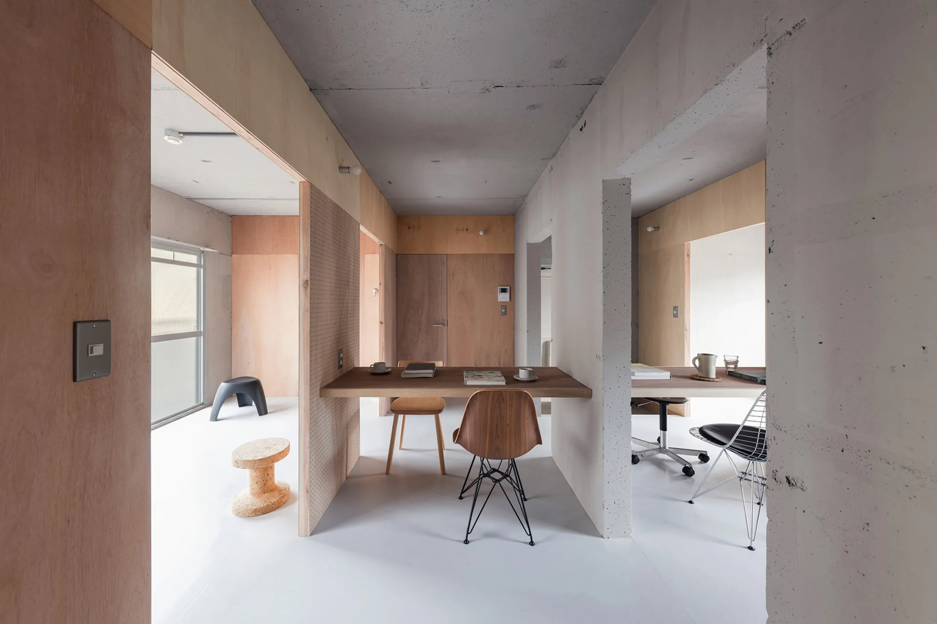 A minimalist home office featuring wooden desks, a pegboard partition, plywood walls, and an exposed concrete ceiling with various modern chairs.