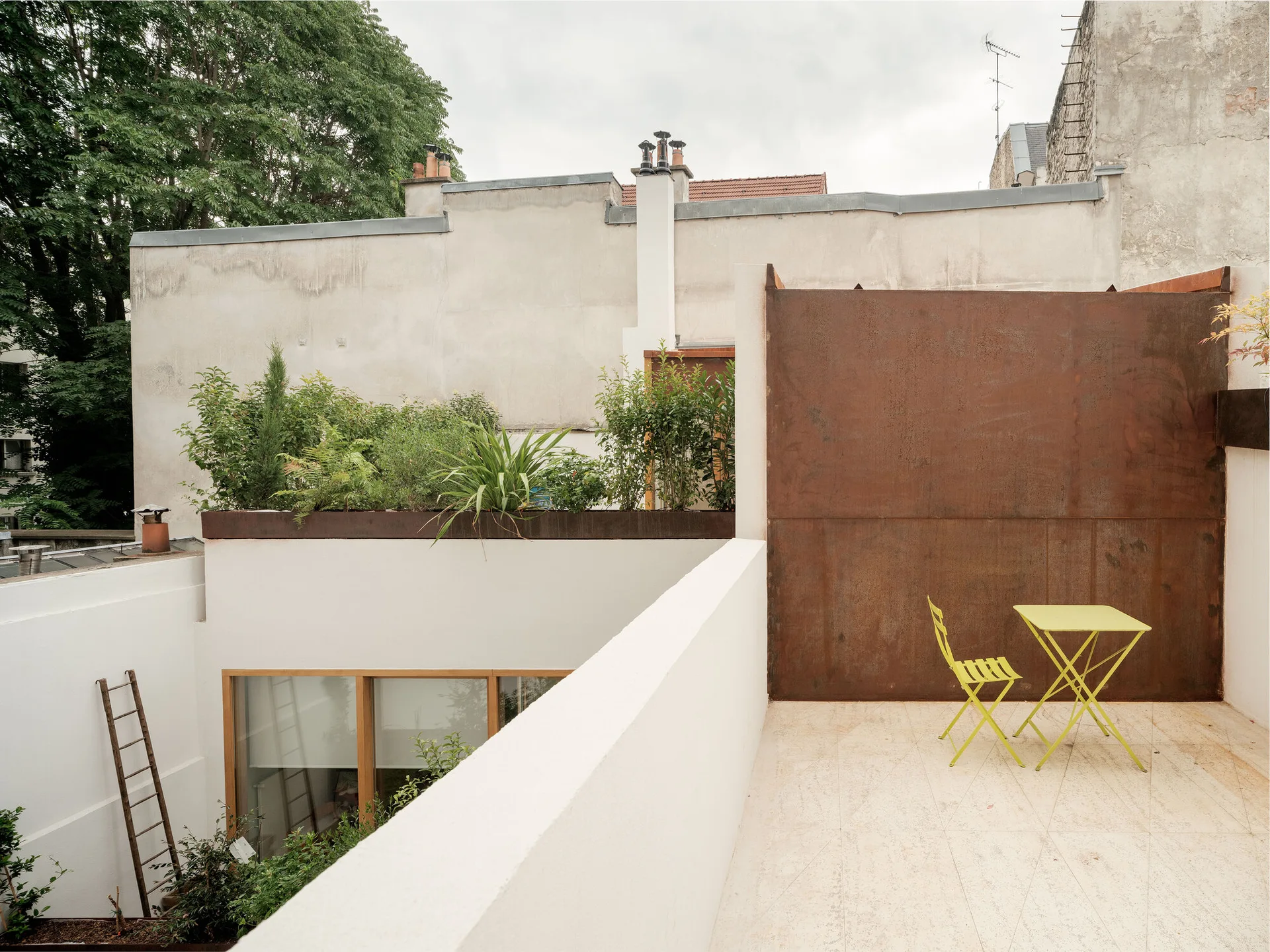 A rooftop terrace features a lime green bistro table and chair set on stone tile, positioned in front of a weathered corten steel wall.