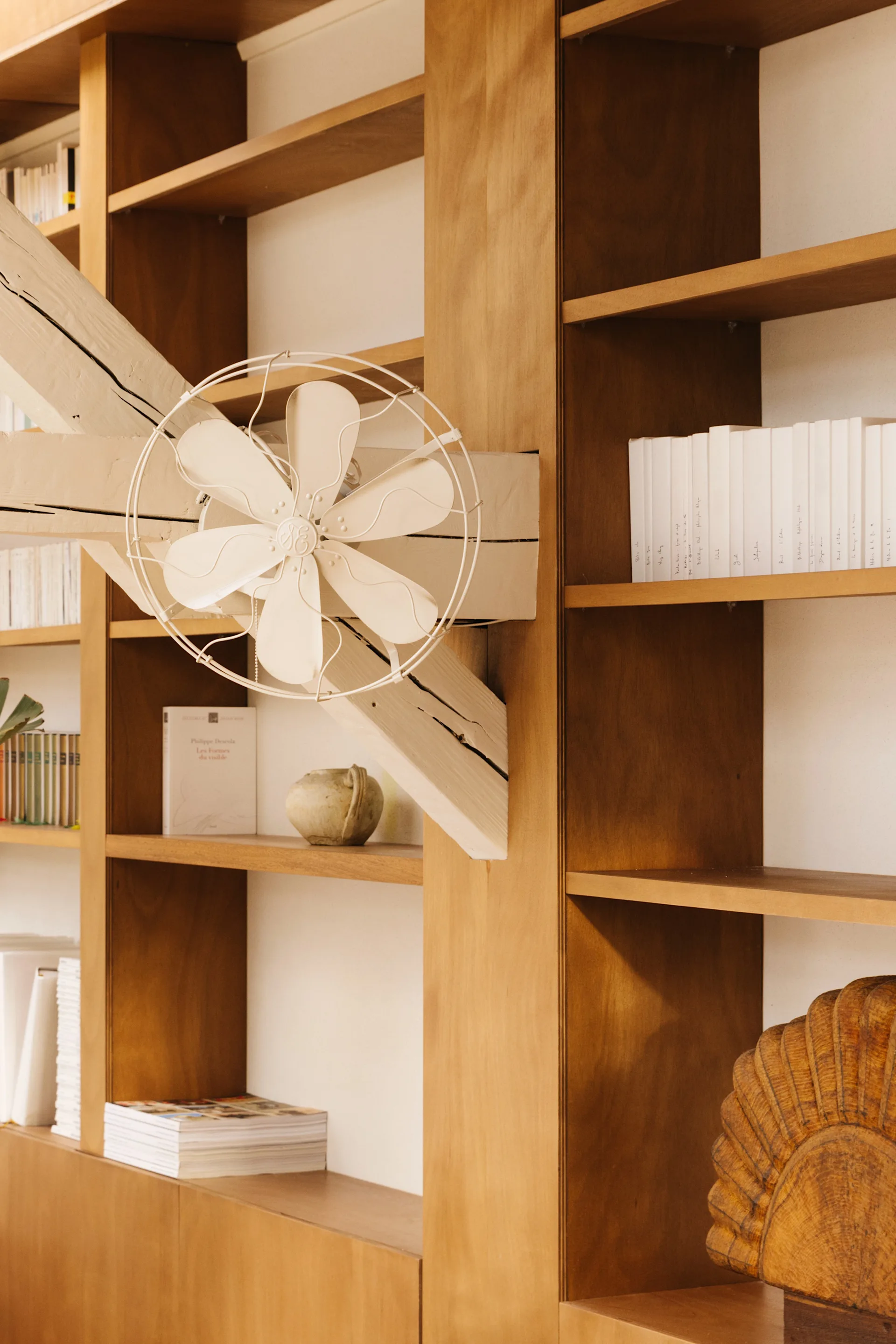 A close-up of a warm wood built-in bookcase featuring a vintage off-white metal fan mounted on an intersecting white structural beam.