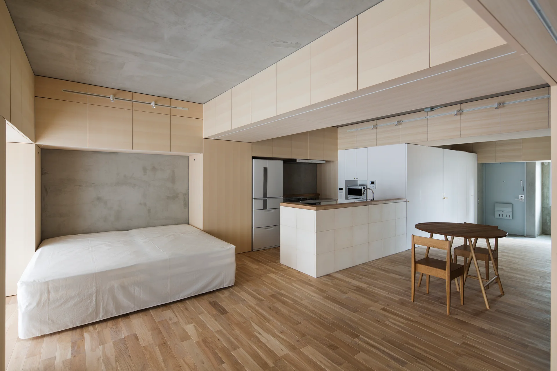 A minimalist studio apartment features a low bed in a wooden niche, light wood overhead cabinets, an exposed concrete ceiling, and a kitchen island.