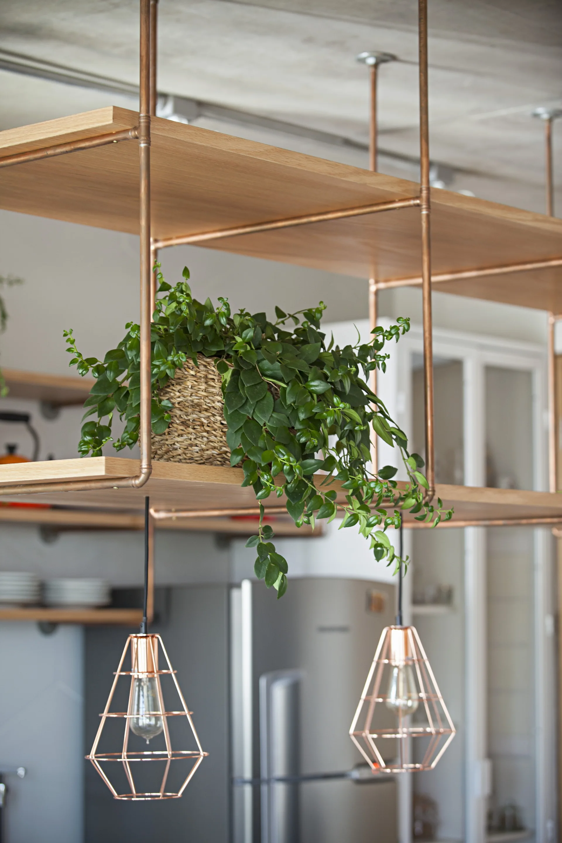 A kitchen scene with suspended wood shelves built with a copper frame and hanging geometric pendant lights.