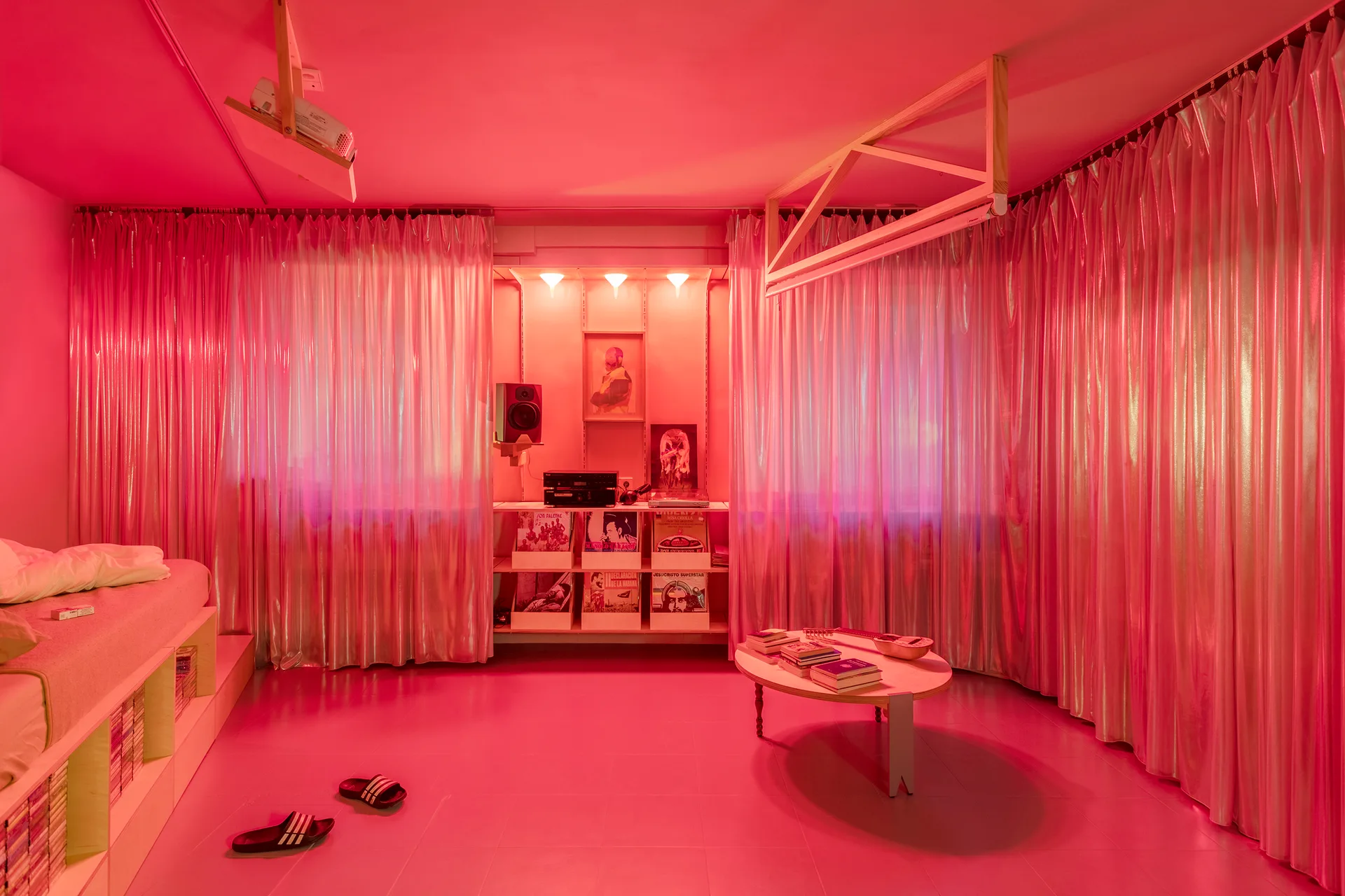 A monochromatic bedroom bathed in pink light featuring metallic curtains, a platform bed with built-in storage, and a low round coffee table.