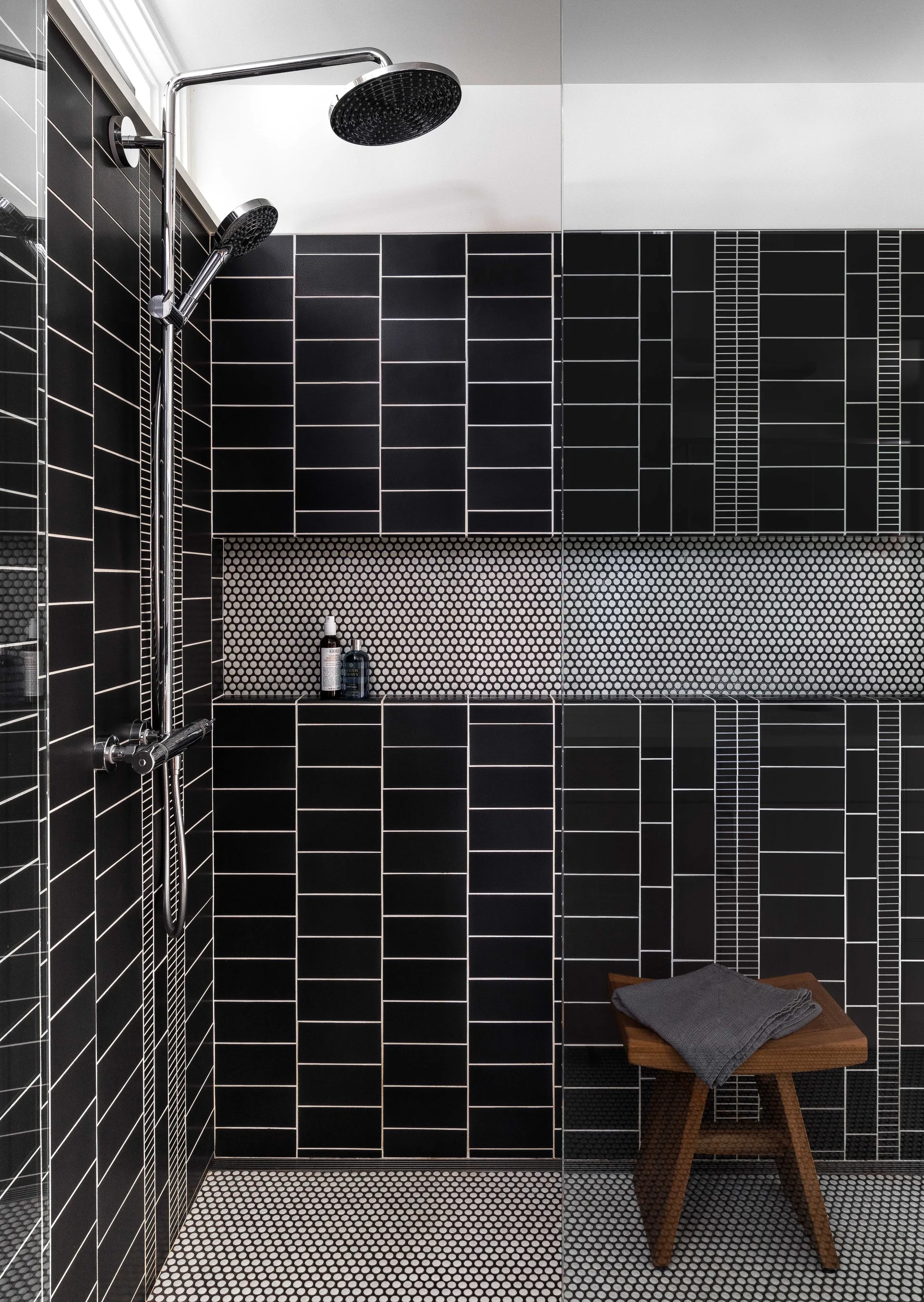 A modern shower features black vertically stacked subway tiles, a recessed niche with penny round mosaic lining, and a chrome rainfall shower head.