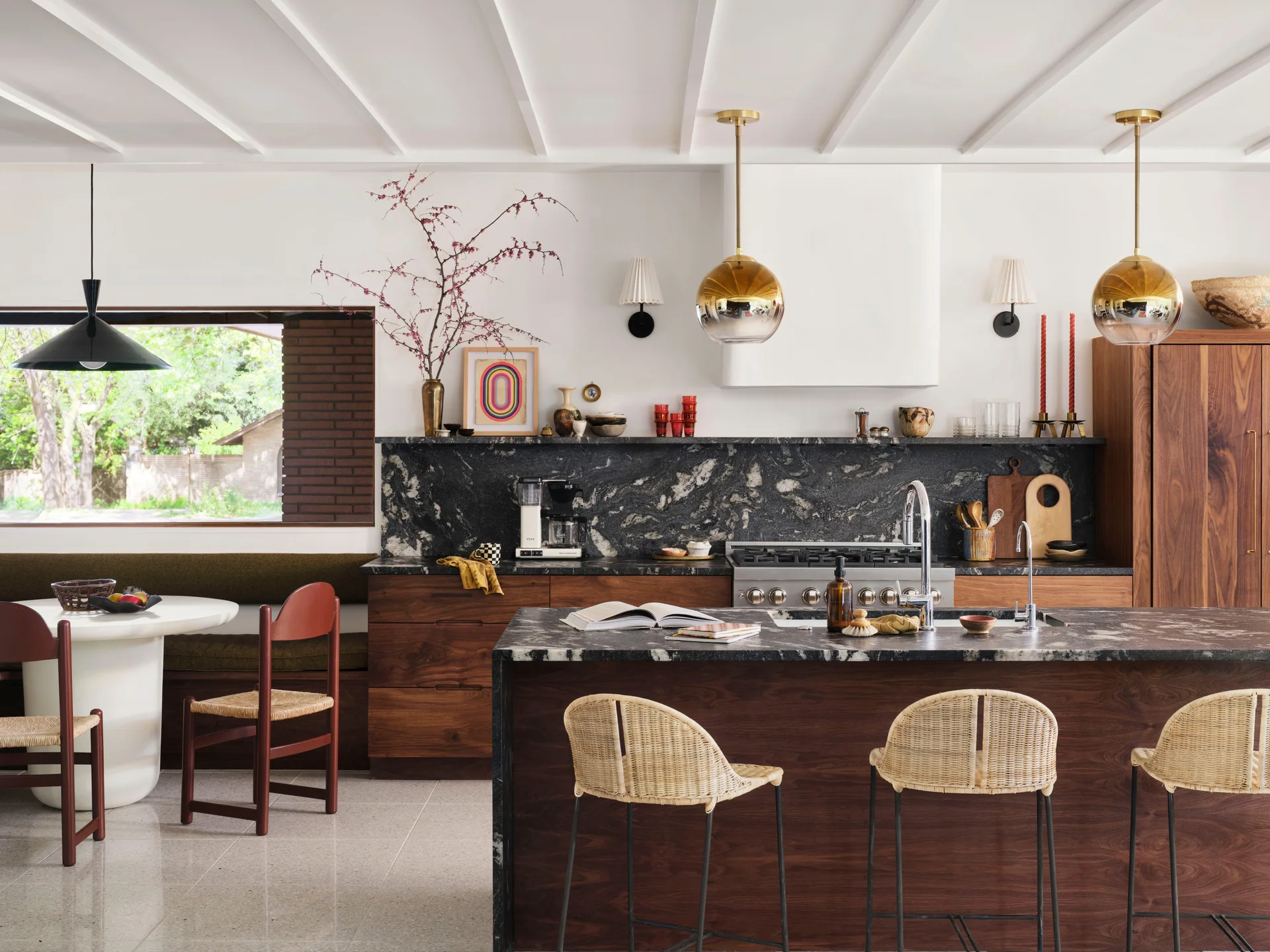 A contemporary kitchen with dark walnut cabinetry, a highly veined black stone island, woven rattan bar stools, and an adjacent dining nook.