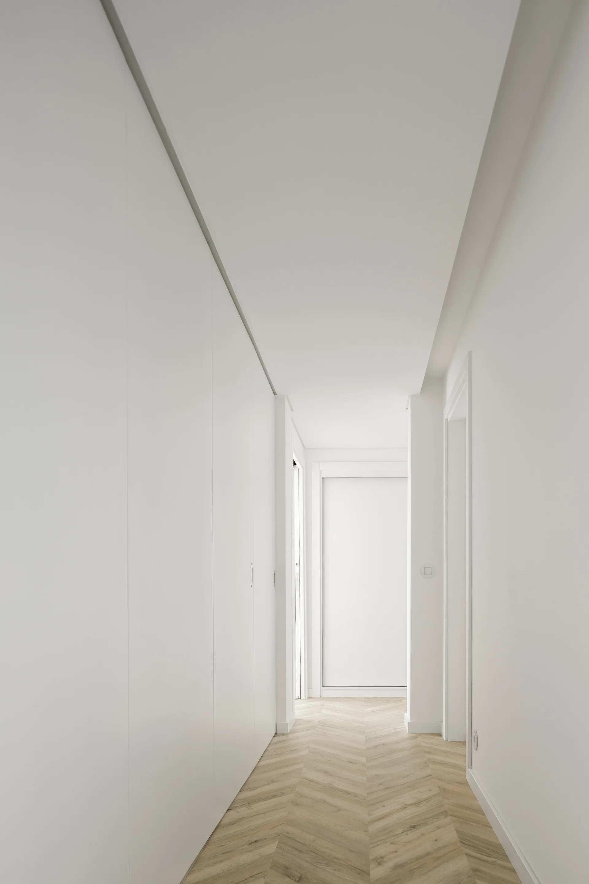 A minimalist white hallway featuring seamless built-in cabinet walls and light wood herringbone flooring.