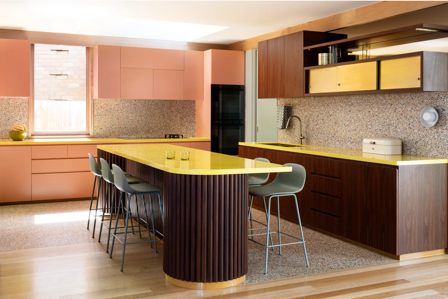 A modern kitchen features a curved fluted wood island with a yellow countertop, peach and walnut cabinets, grey stools, and terrazzo flooring.