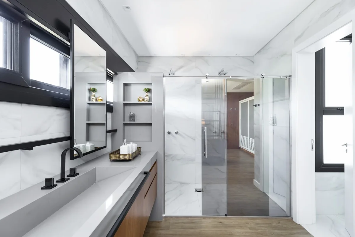 A modern bathroom features a long grey vanity with an integrated sink, marble-look tiled walls, and a sliding glass shower door.