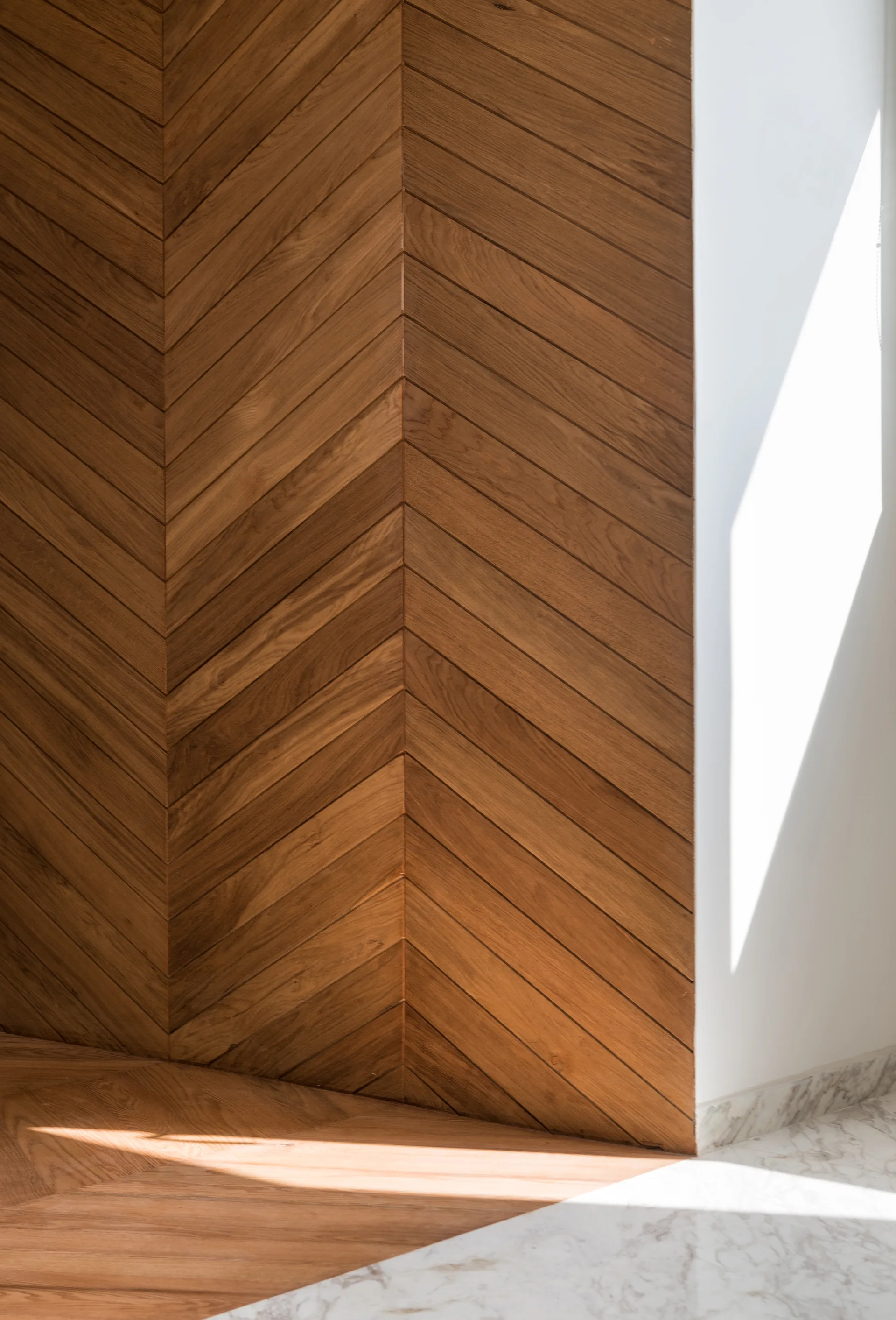 A close-up of a wall corner featuring warm brown wood paneling in a chevron pattern next to a white wall and white veined marble tile flooring.
