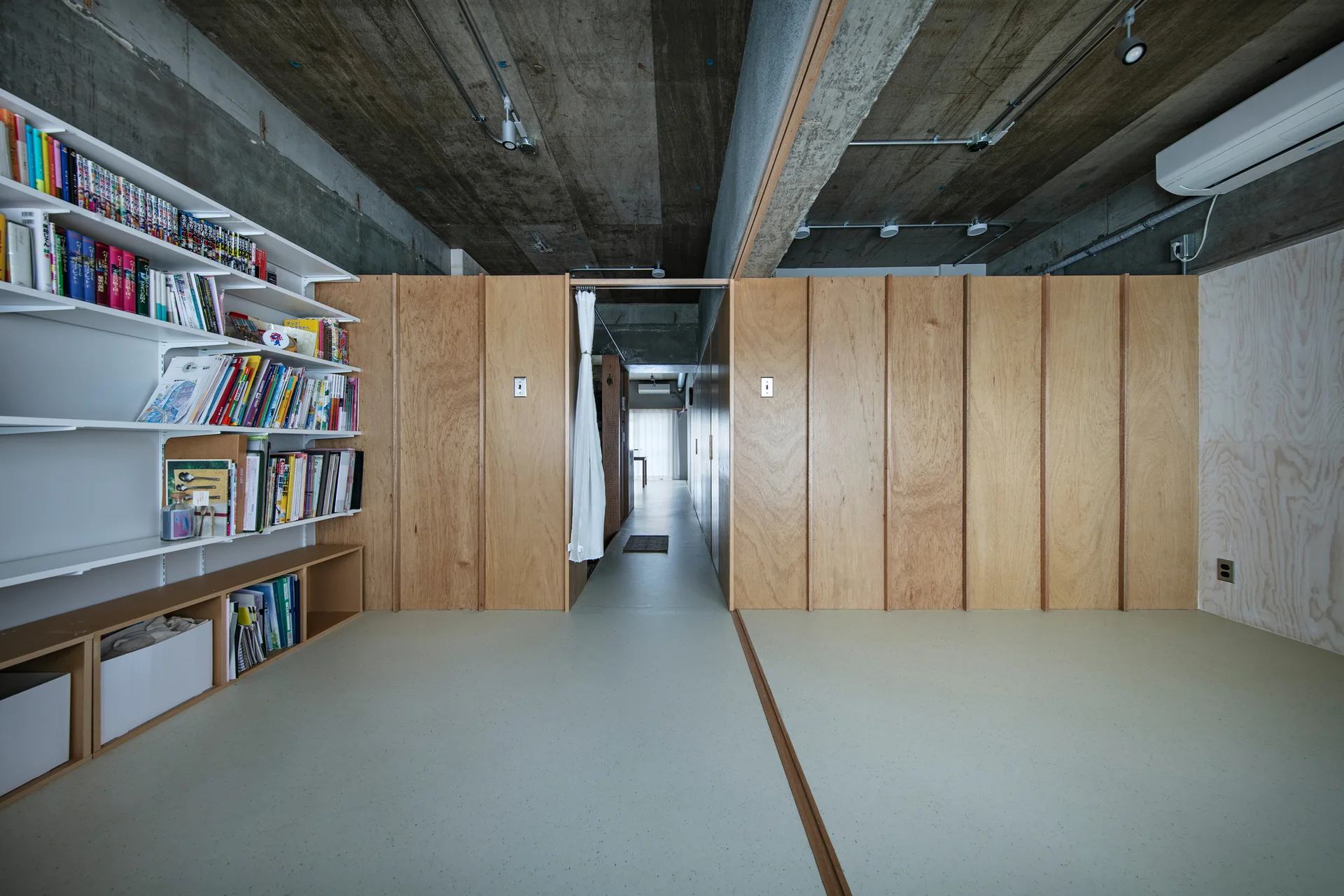 An interior hallway features light-colored plywood partition walls, a white wall-mounted bookshelf filled with books, and an unfinished concrete ceiling.