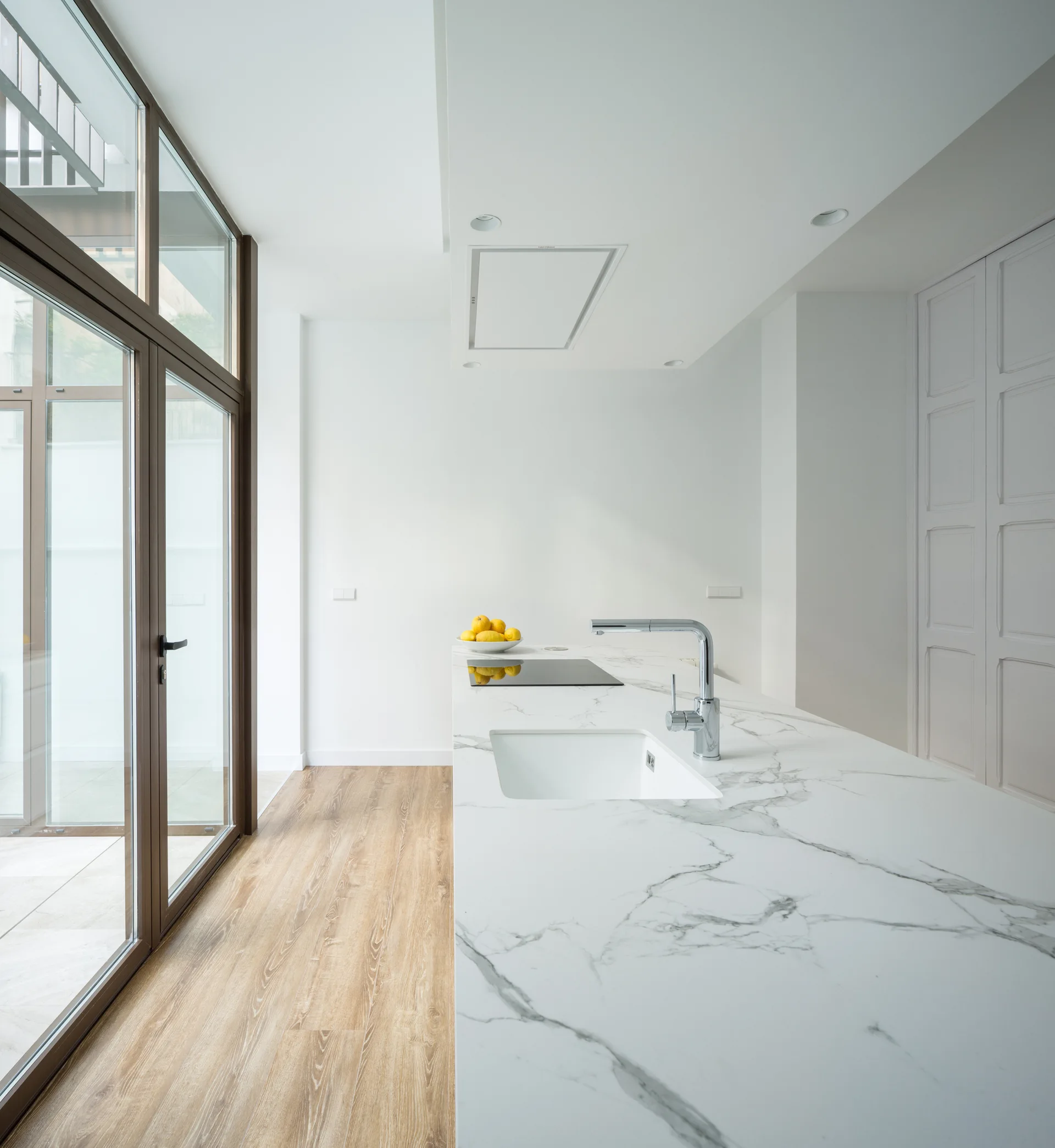 A minimalist kitchen features a white marble-patterned island with an integrated sink alongside light wood floors and large glass doors.