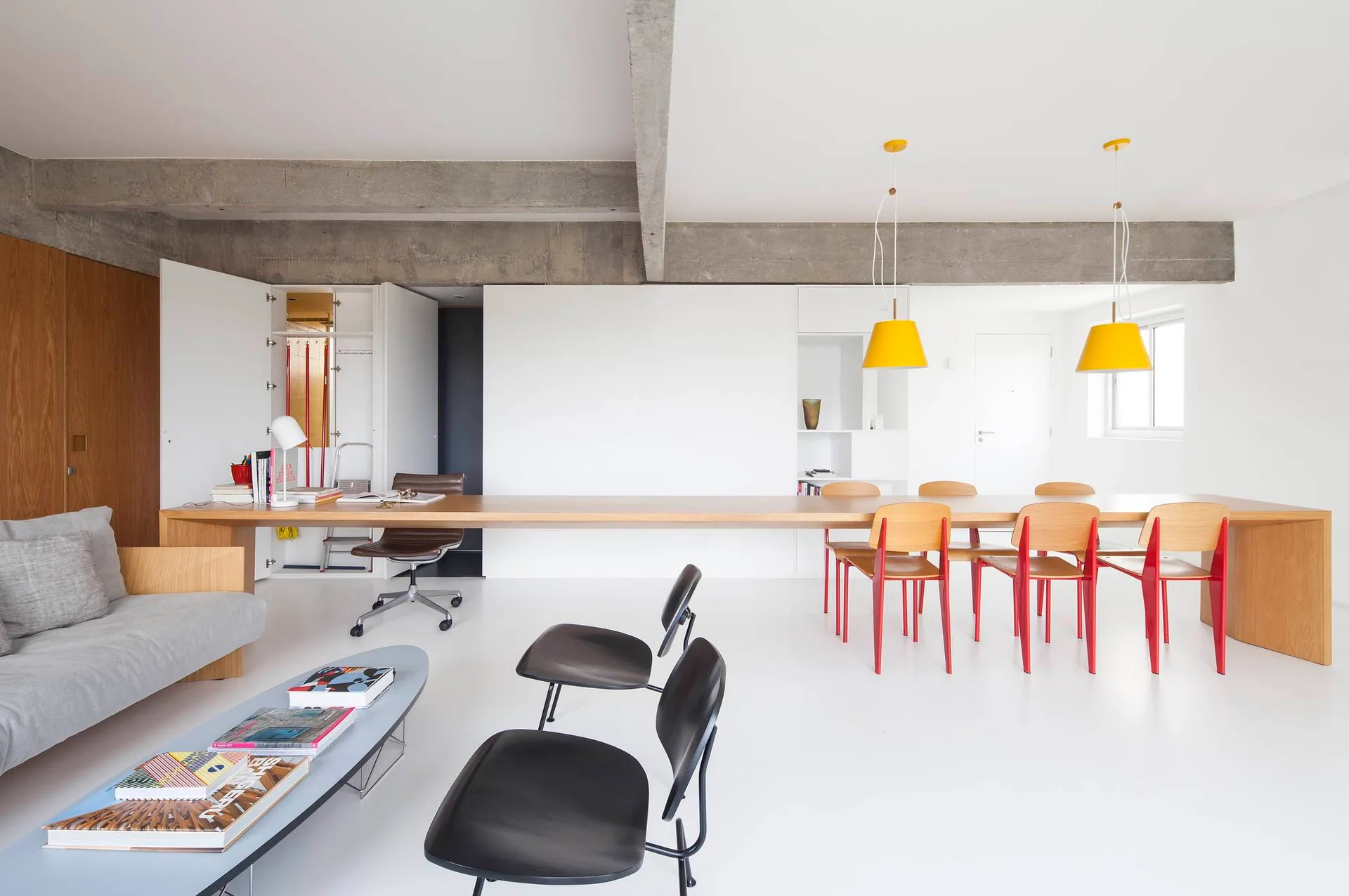 A minimalist white room features a long wooden table serving as both a desk and a dining table, beneath an exposed concrete ceiling beam.