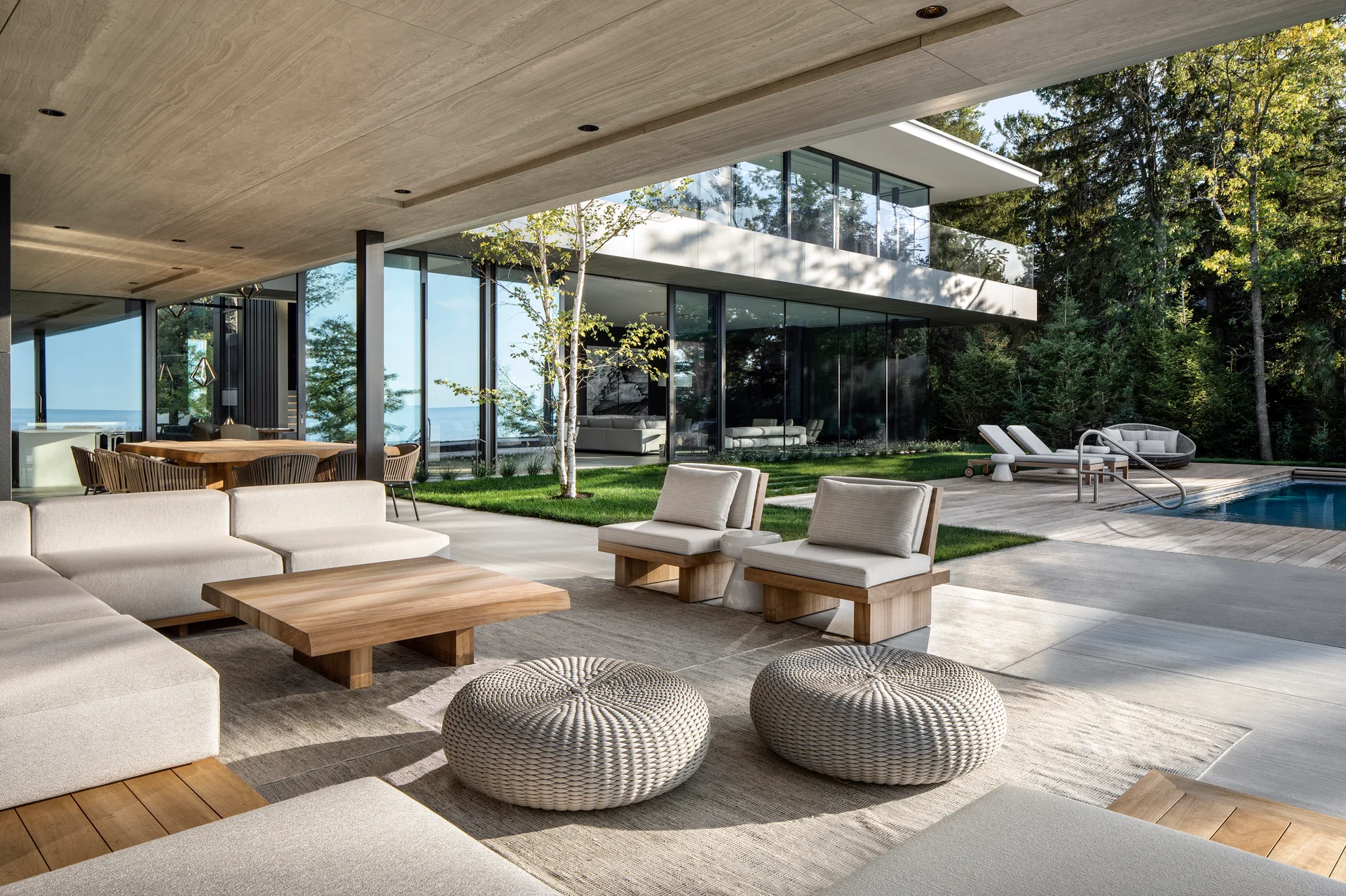 A modern covered patio features a large cream sectional, a low wood coffee table, and woven pouffes beneath a wood plank ceiling next to a pool.