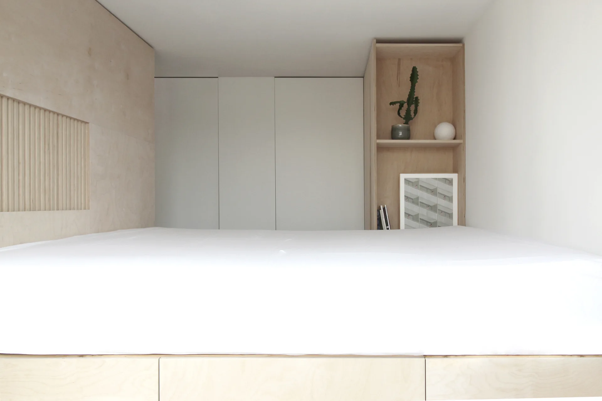 A minimalist bedroom featuring a platform bed integrated with light plywood cabinetry, built-in shelving holding a cactus, and a white wardrobe.