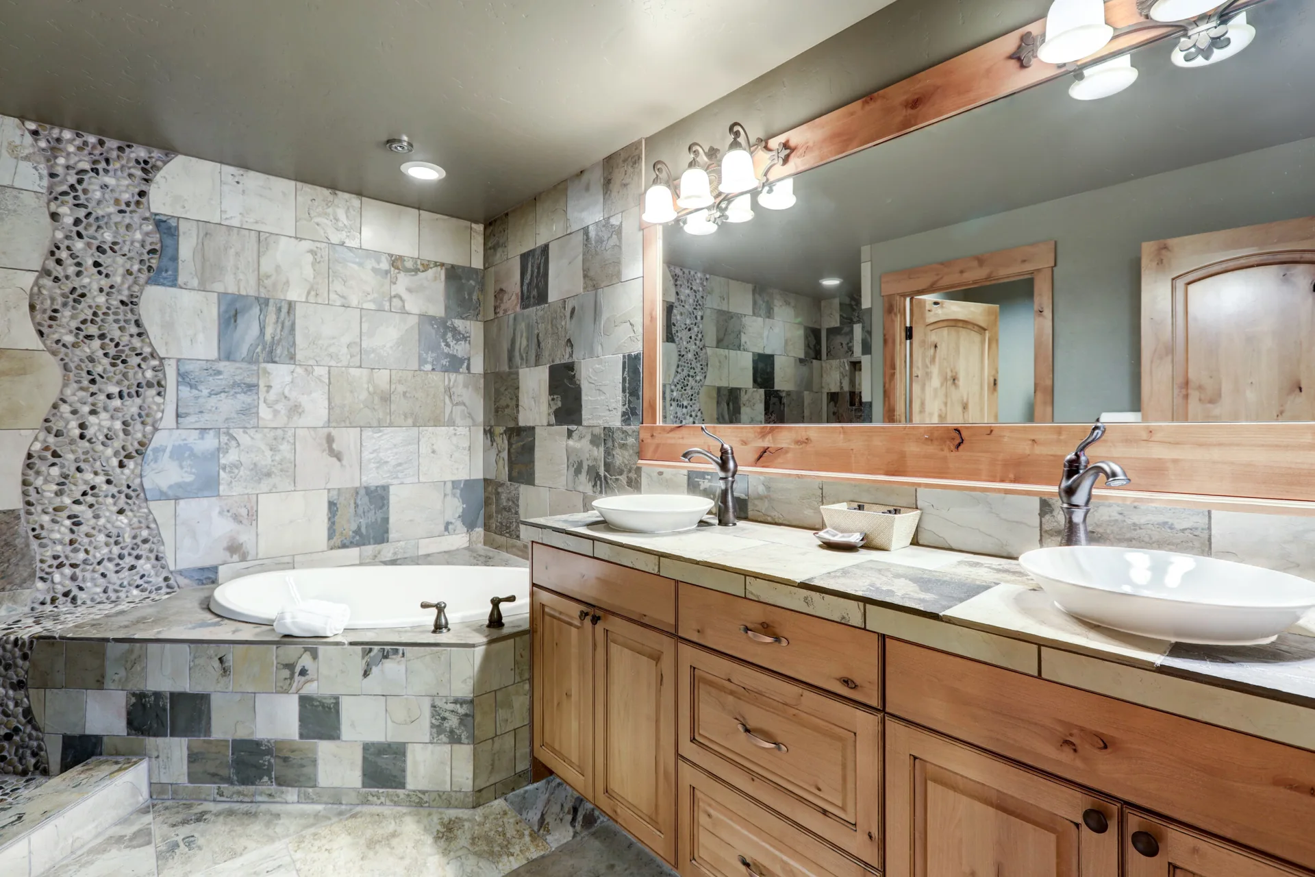 A rustic bathroom features a wooden double vanity with white vessel sinks, a stone tile backsplash, and a built-in corner bathtub.