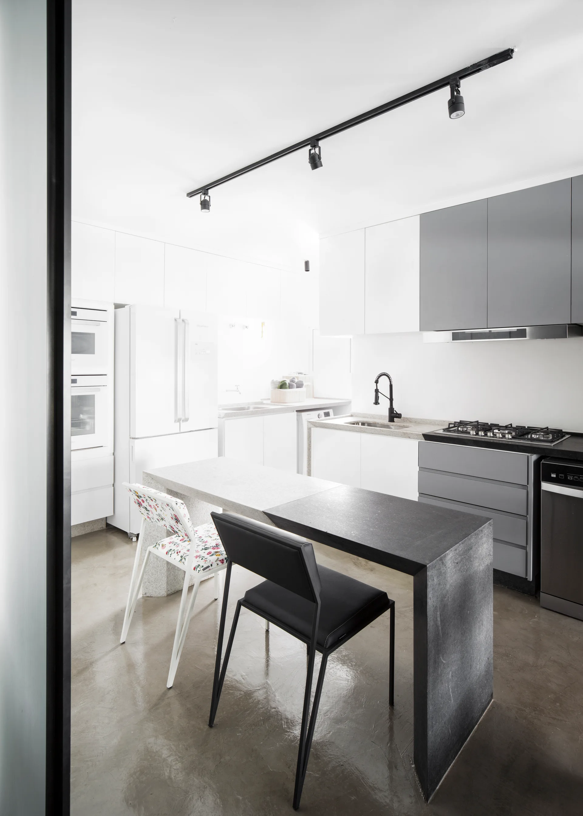 A minimalist kitchen features a split-tone waterfall island with two chairs, white cabinetry, and a polished concrete floor.