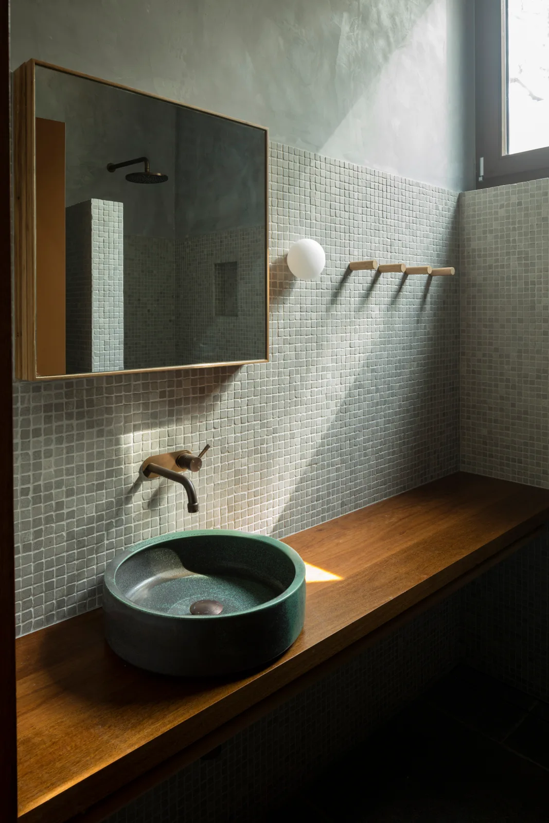 A green vessel sink sits on a floating wood counter against a wall of pale green mosaic tiles with a square wooden-framed mirror.