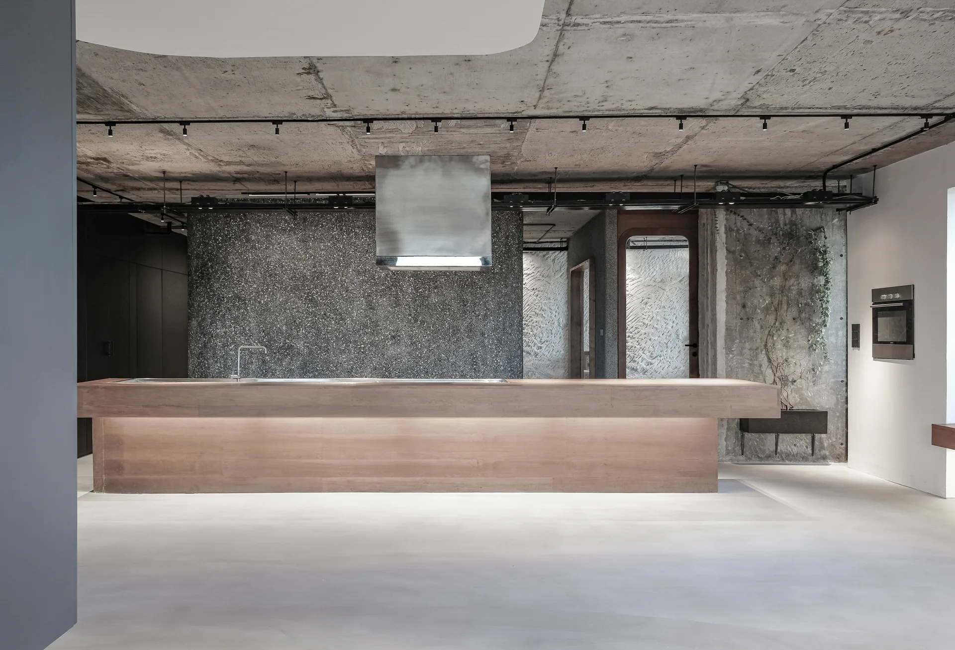 A minimalist kitchen features a long wooden island, a dark textured accent wall, and a raw exposed concrete ceiling with track lighting.