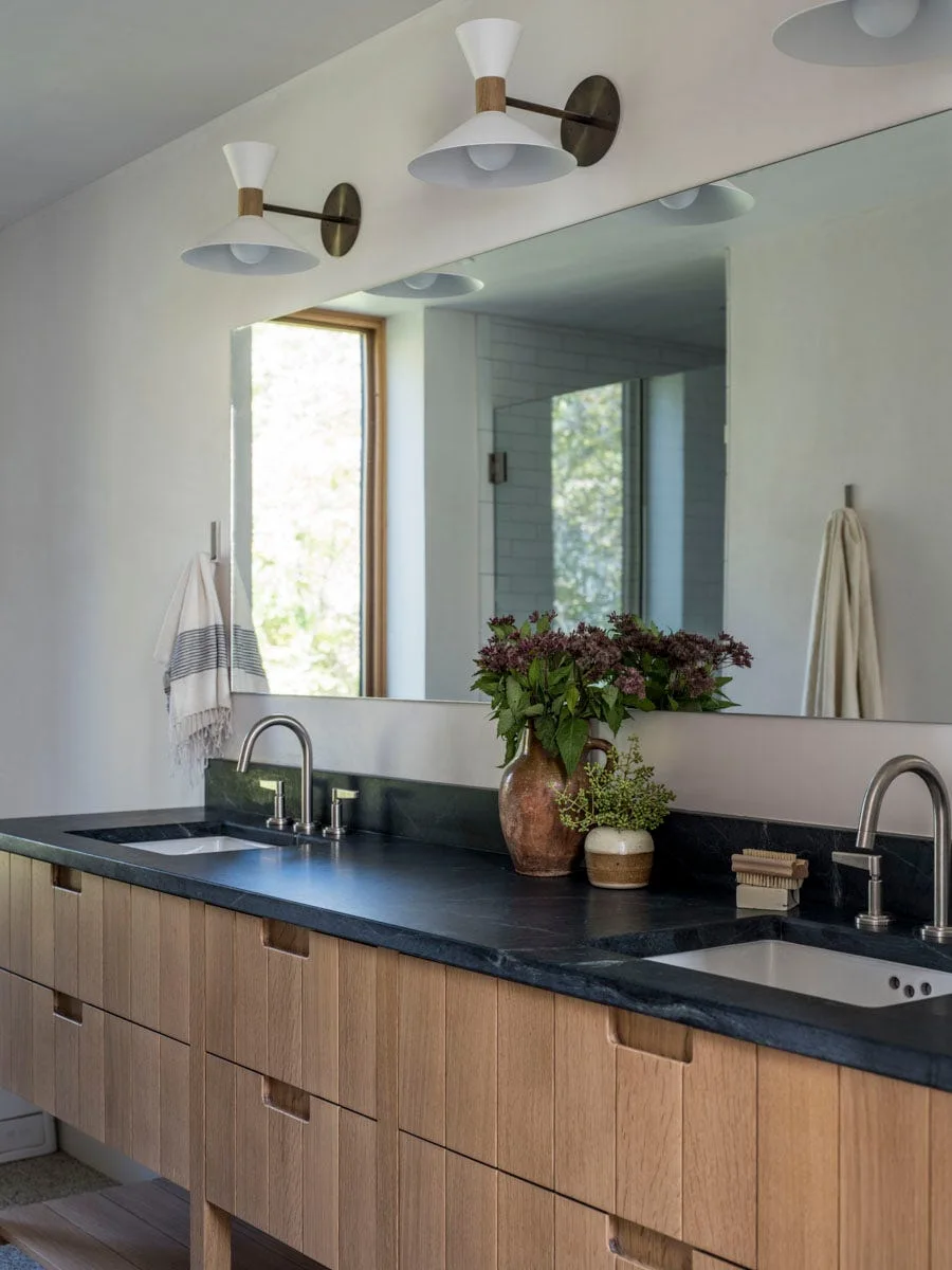 A modern bathroom double vanity features light wood vertical plank cabinetry, a dark stone countertop, two chrome faucets, and a large wall mirror.