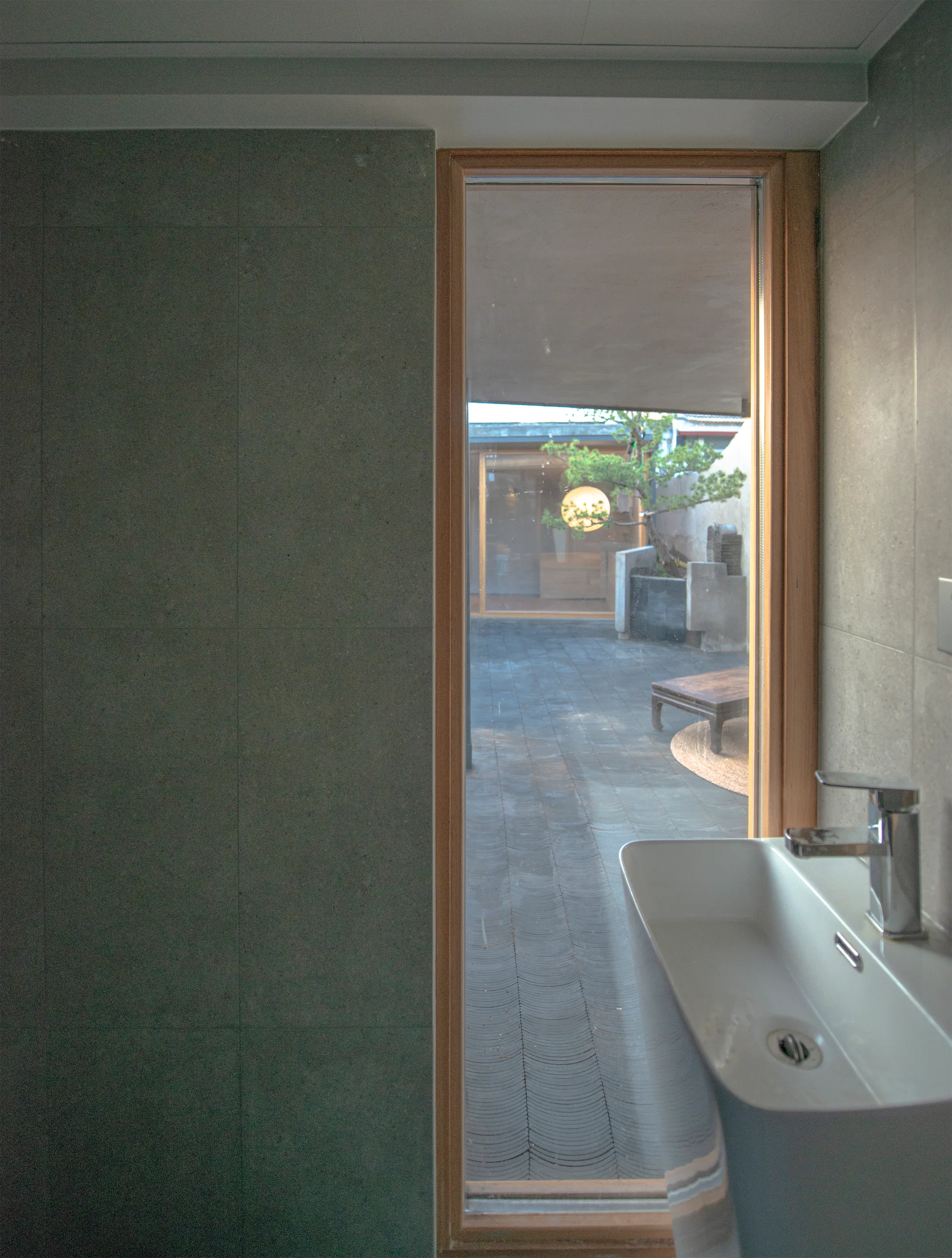 A modern bathroom with a white wall-mounted sink, large grey tile walls, and a tall wood-framed window overlooking an outdoor stone courtyard.