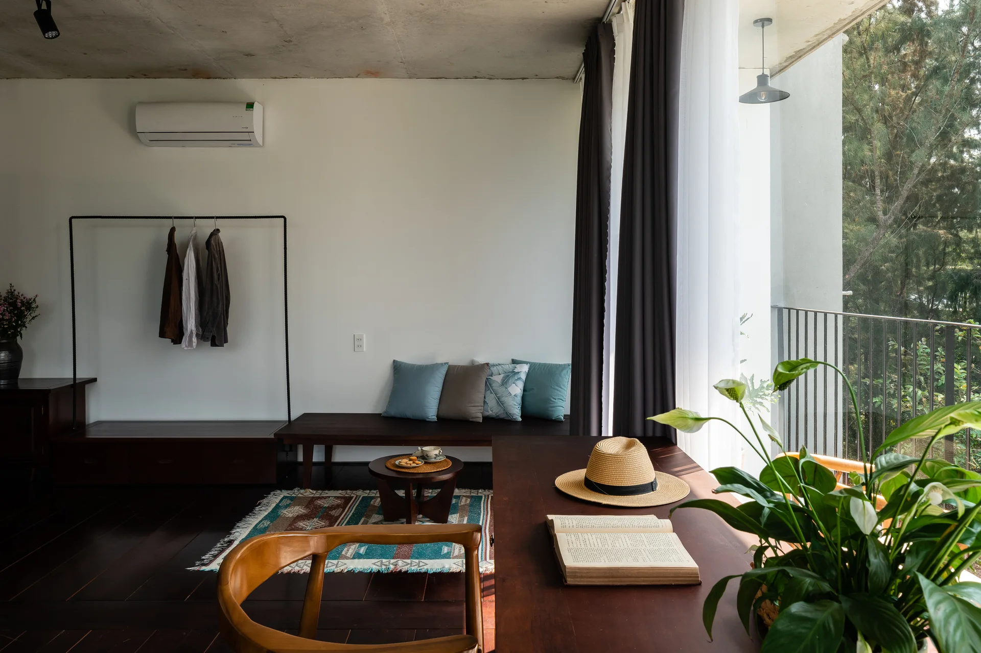 A minimalist bedroom featuring a dark wood desk, a low wooden bench with blue pillows, a simple clothing rack, and an exposed concrete ceiling.