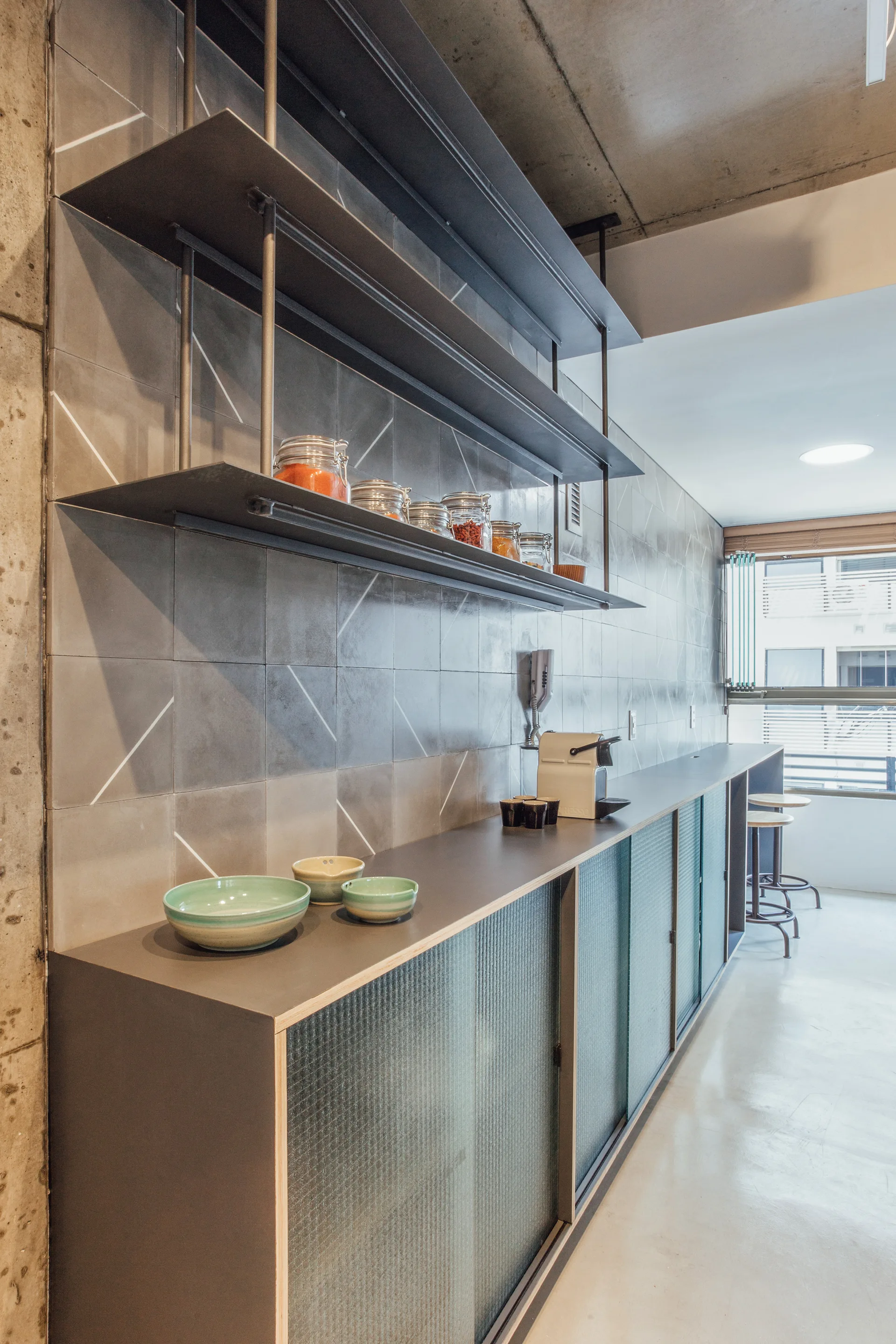 A long grey storage cabinet with frosted glass sliding doors stands beneath steel shelves on a grey tiled wall under a concrete ceiling.