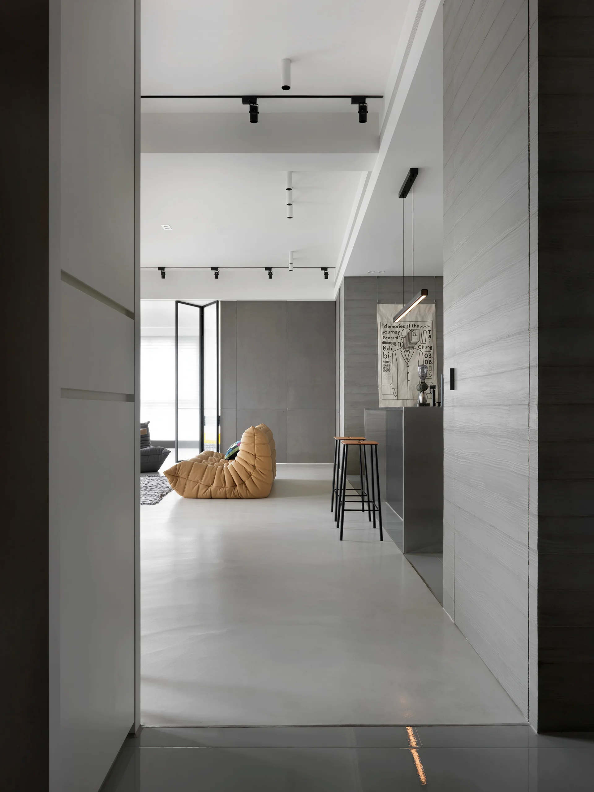 An open-concept minimalist room features a tan tufted lounge chair, a sleek metal bar counter with stools, and large grey wall panels.
