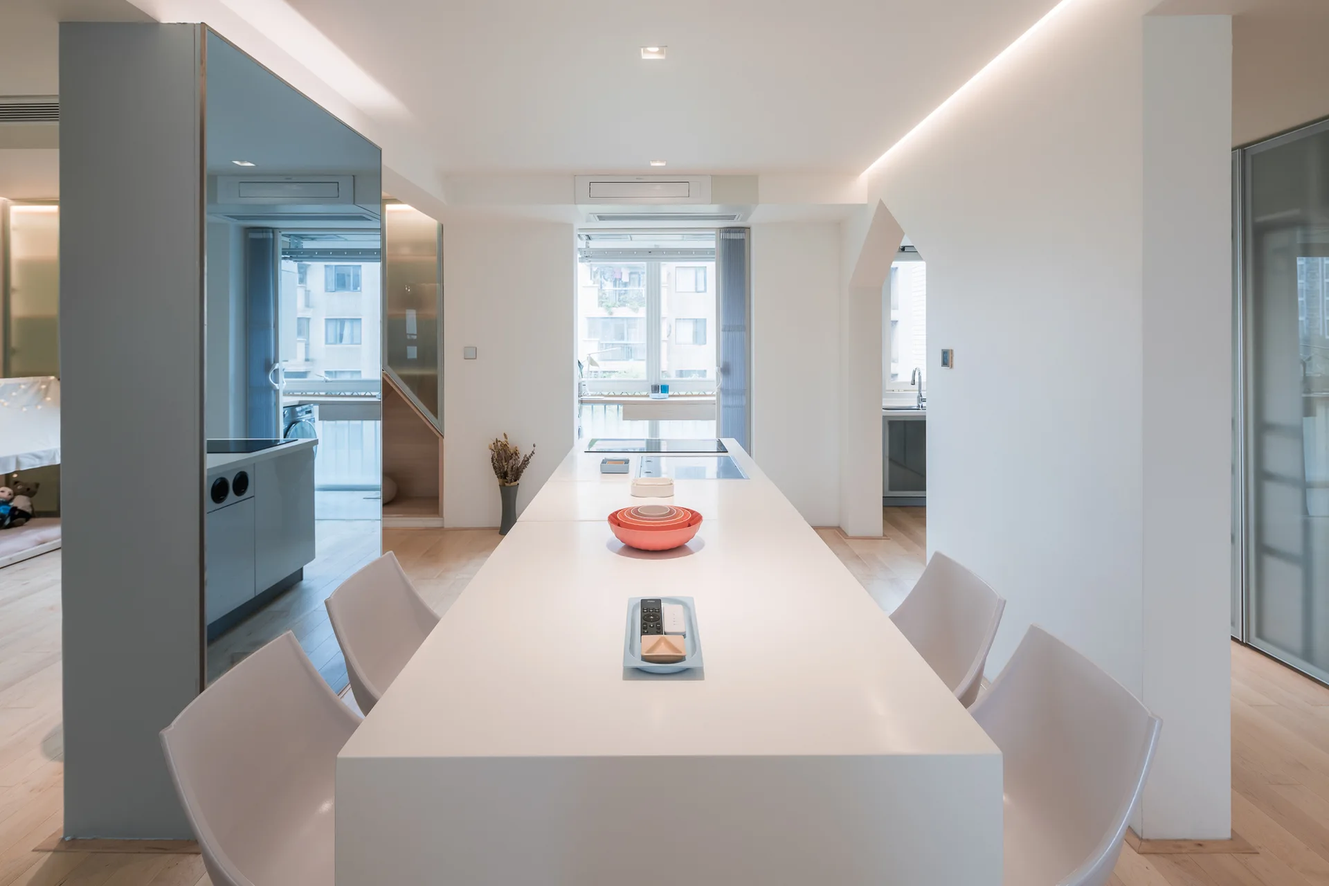 A minimalist kitchen features a long white central island with matching chairs, light wood flooring, and a unique triangular archway.