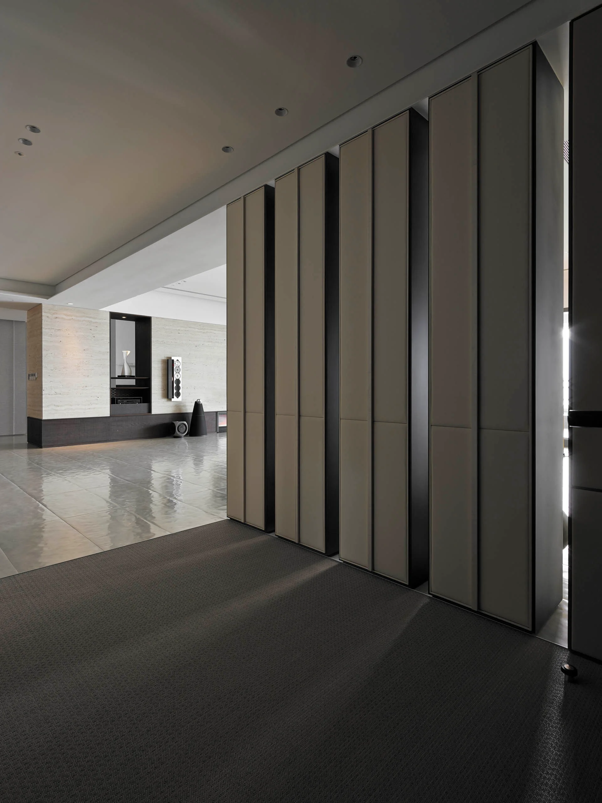 Tall vertical greige cabinetry stands beside a polished tile floor and a horizontal beige stone feature wall in a minimalist interior.