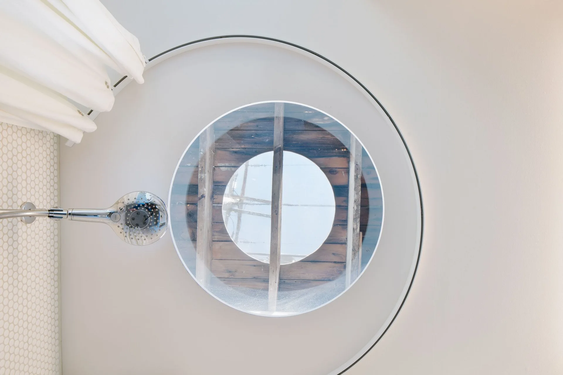A low-angle view of a white bathroom ceiling featuring a circular skylight, a chrome shower head, a curved curtain track, and white mosaic wall tiles.