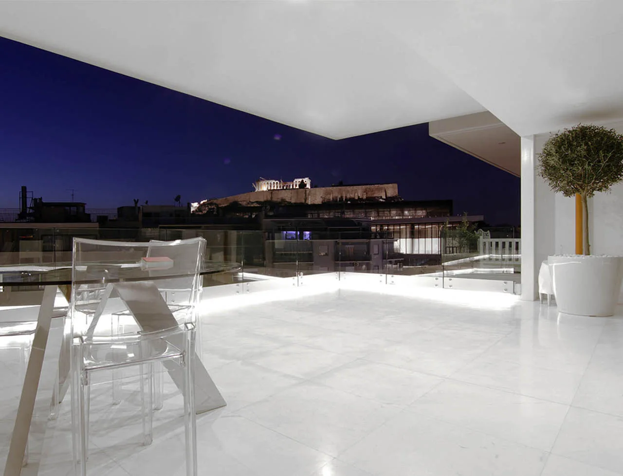 A white rooftop terrace at night features transparent acrylic dining chairs, a glass railing, and a view of the illuminated Acropolis.