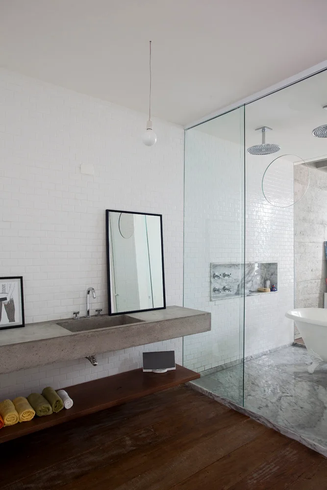 A modern bathroom with a floating concrete vanity, a low wood storage shelf, and a glass-enclosed shower featuring white subway tiles.