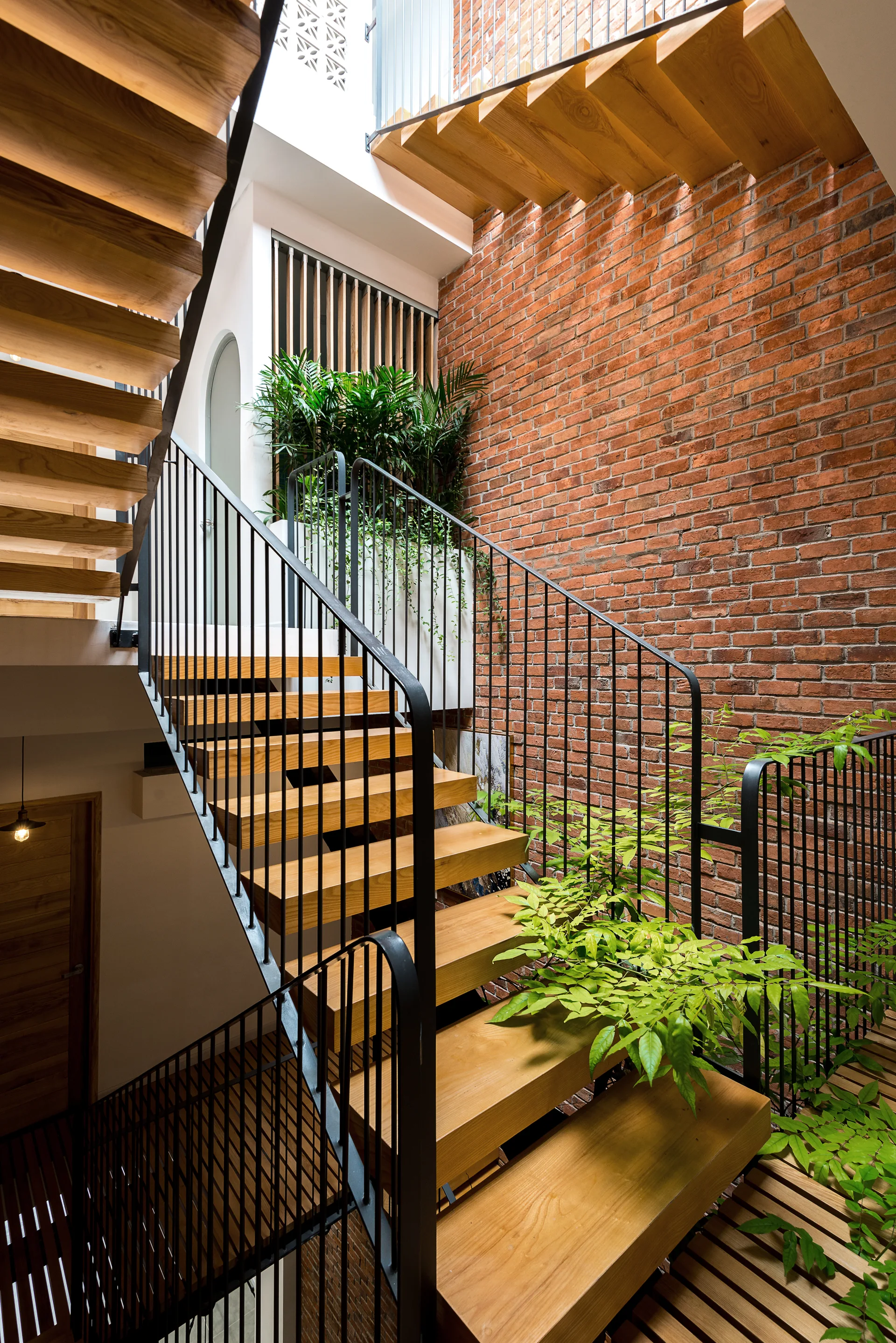 A multi-level floating wooden staircase features thin black metal railings against a high exposed red brick wall with lush green indoor plants.