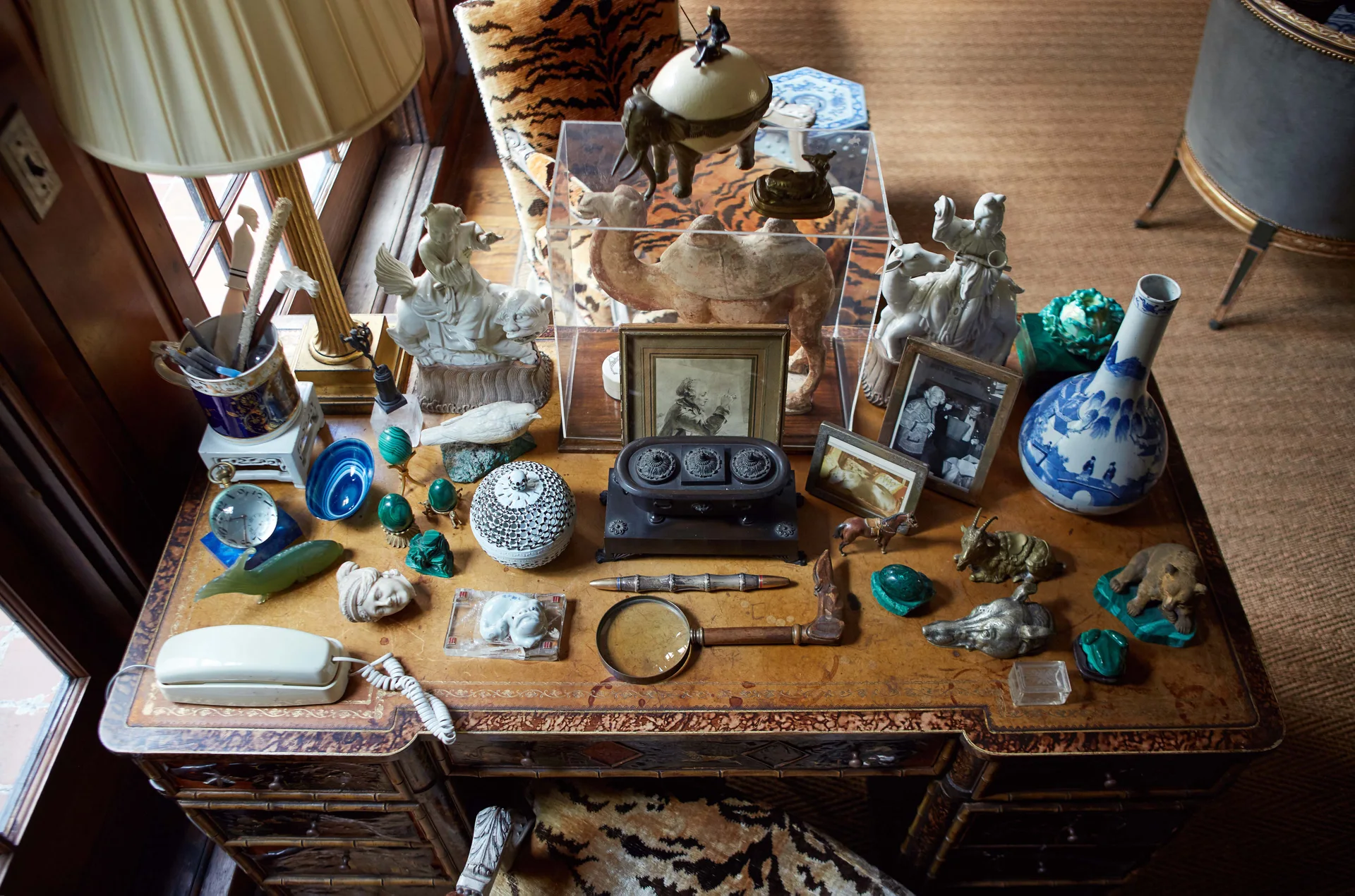 A high-angle view of a carved wooden desk covered with an extensive collection of ceramic figurines, vintage photographs, and a magnifying glass.