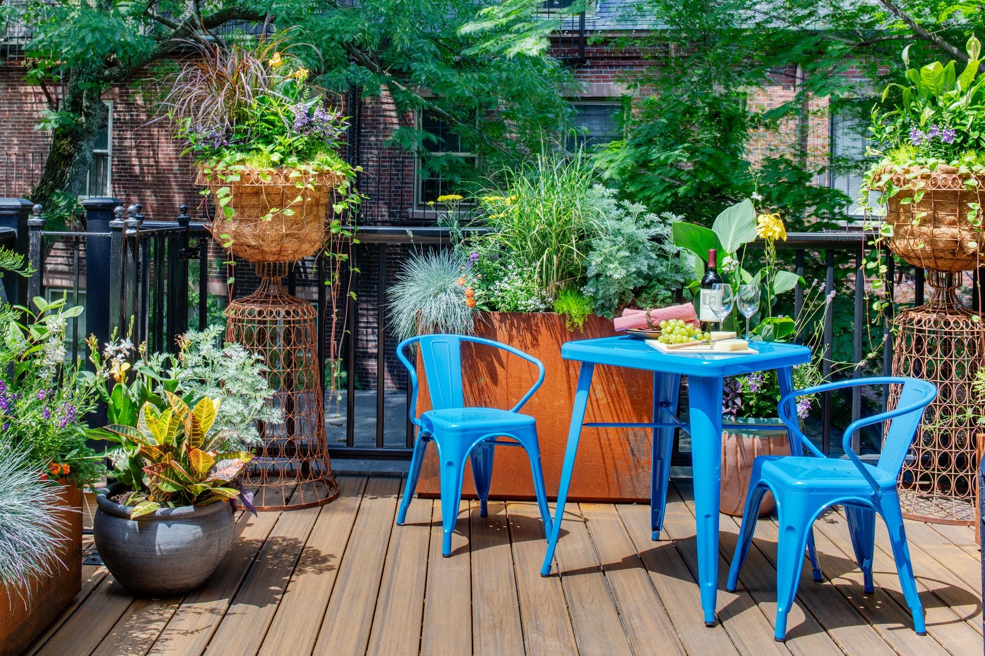 A blue metal bistro table and chairs sit on a wooden patio deck surrounded by potted green plants and wire pedestal planters.