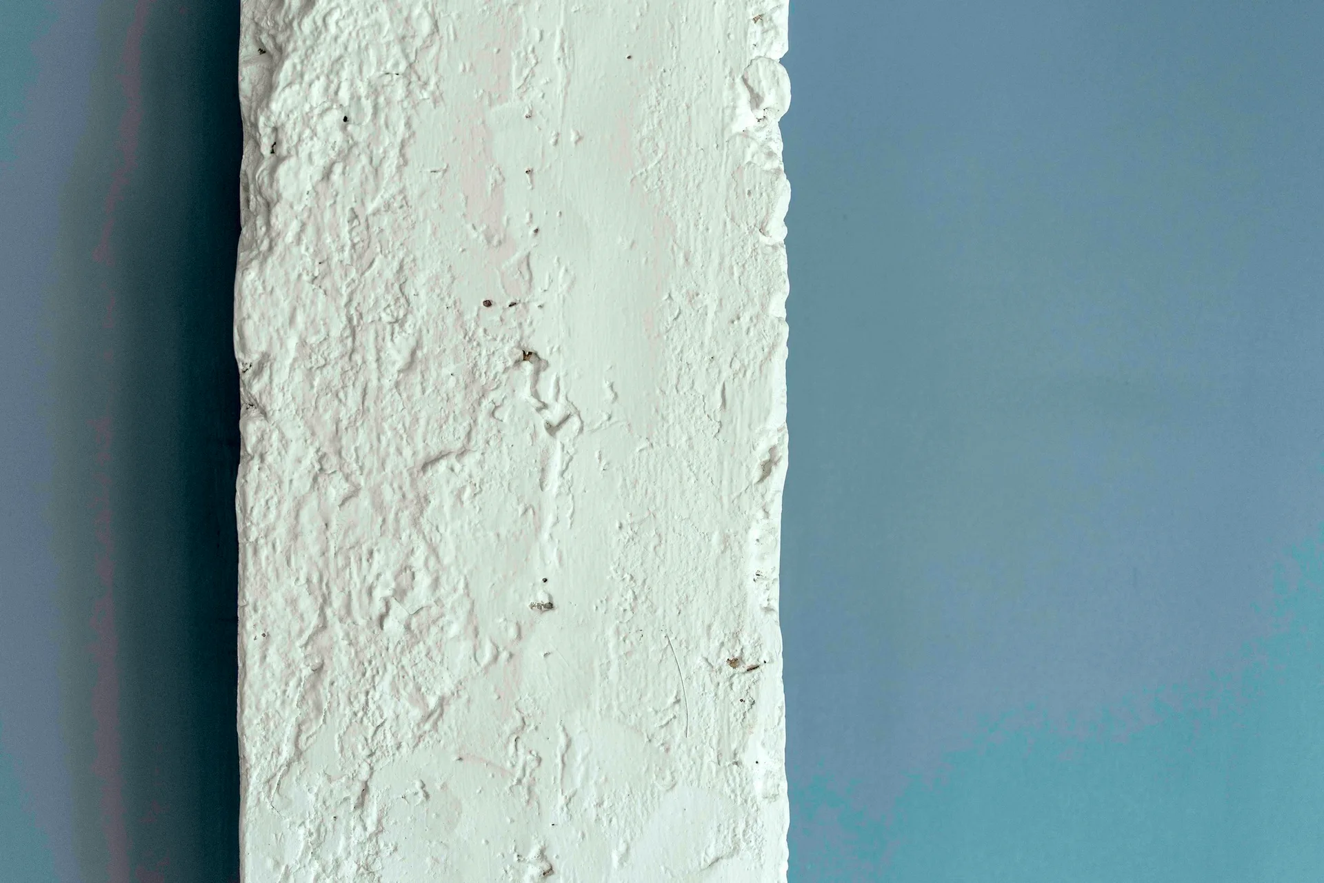 A close-up of a heavily textured white painted masonry column set against a smooth matte grey wall background.