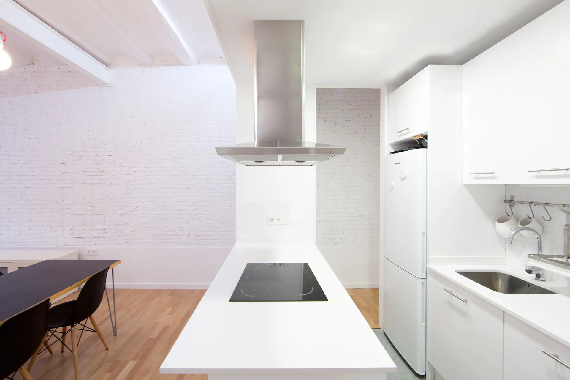 A modern white kitchen featuring a central island with a built-in cooktop, a stainless steel range hood, and a white painted brick wall.