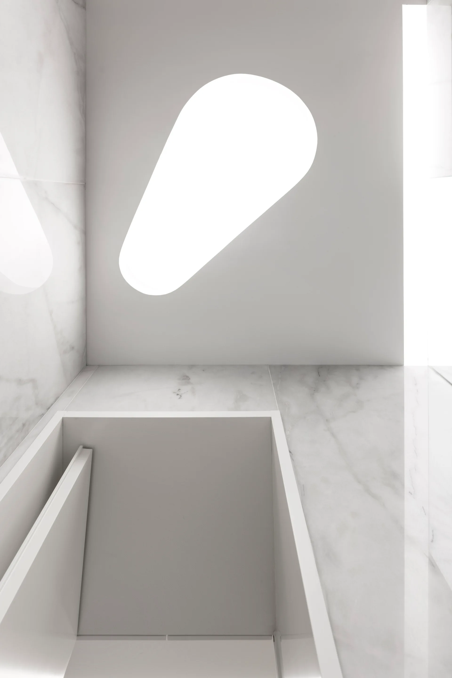 A minimalist bathroom featuring a rectangular recessed sink integrated into a white veined marble countertop beneath a capsule-shaped ceiling skylight.