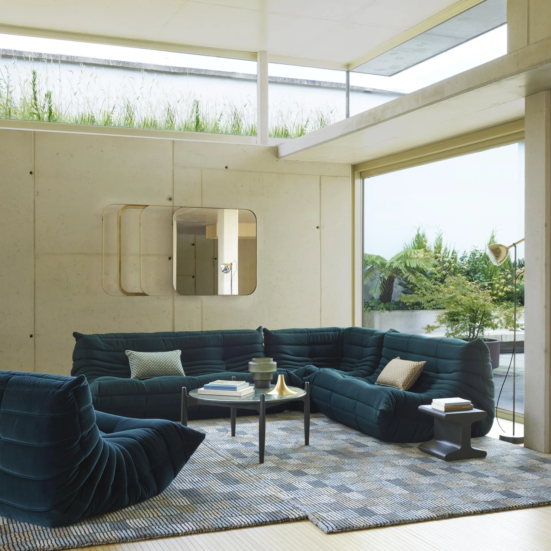 A modern living room featuring a dark green tufted sectional sofa, a matching armchair, a patterned rug, and smooth concrete walls.