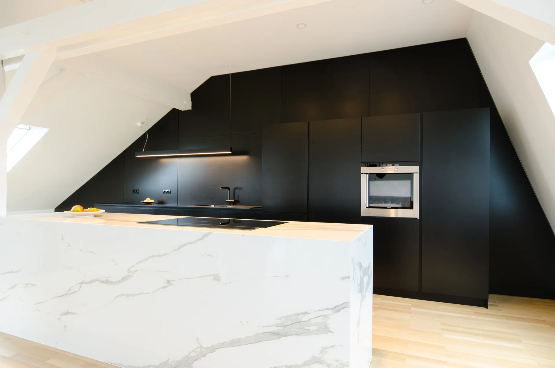 Minimalist attic kitchen with a large marble island, matte black cabinetry, and a sloped white ceiling.