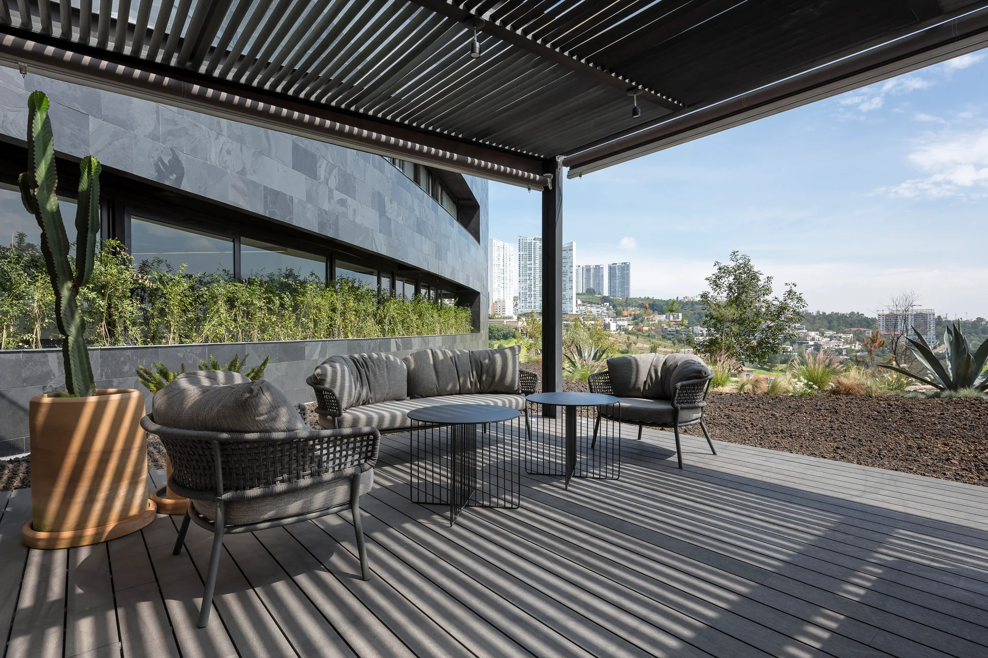 Gray woven outdoor seating sits on a composite deck under a black slatted pergola overlooking a distant city skyline.