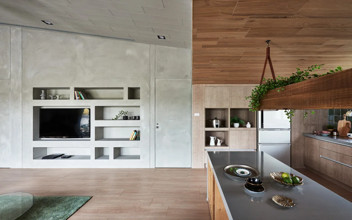 An open-concept interior featuring a concrete wall with built-in shelves on the left and a wood-detailed kitchen with an island on the right.