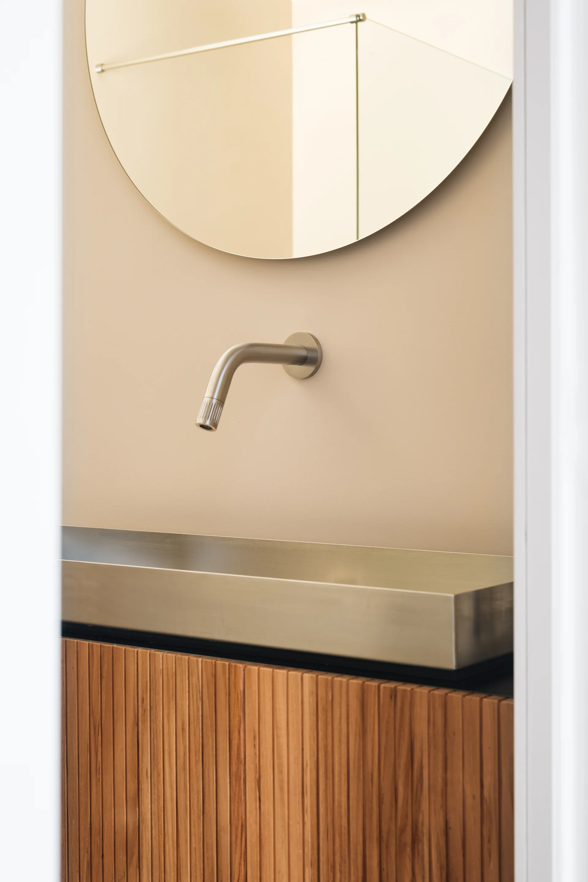 A modern bathroom vanity features a fluted wood cabinet, a shallow metal trough sink, a wall-mounted faucet, and a round mirror against a beige wall.