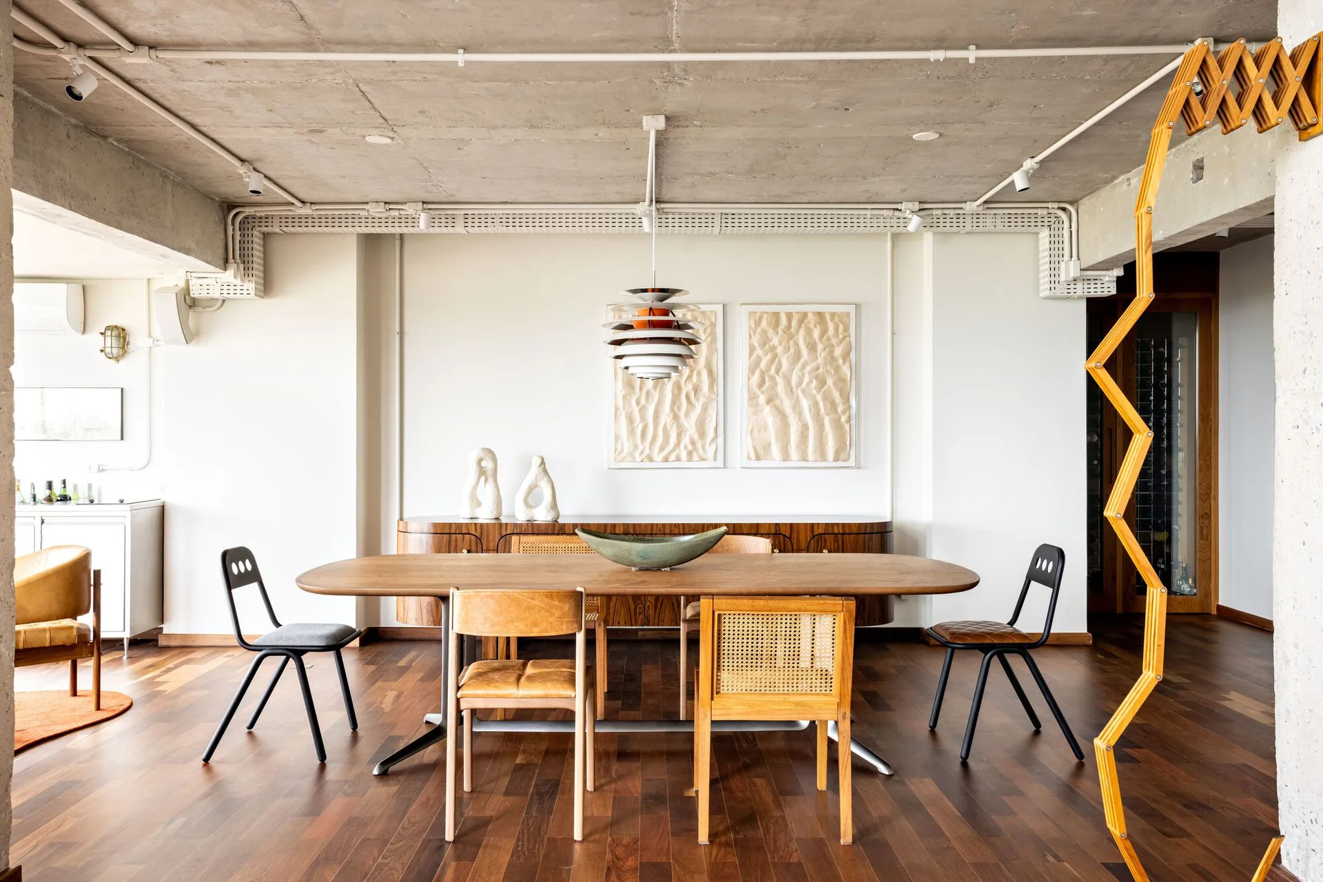A modern dining room features an oval wooden table with mixed chairs, set against a curved wood sideboard beneath an exposed concrete ceiling.