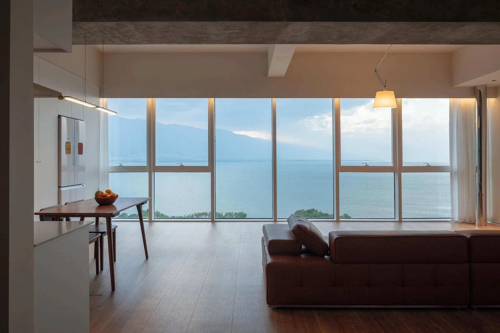 A modern open-concept living area features a brown leather sofa, a wooden dining table, and large windows overlooking a lake and mountains.