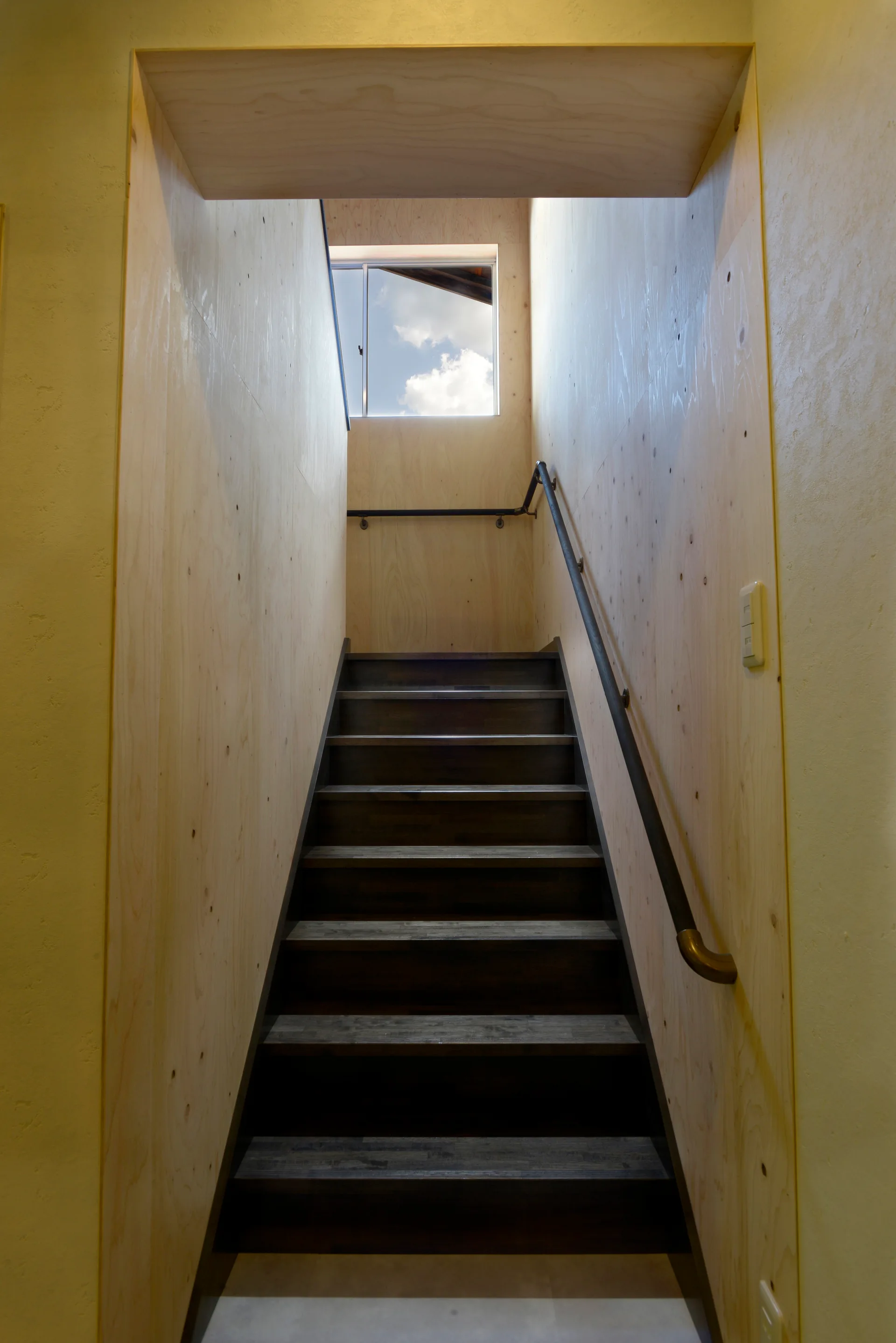 Dark wooden stairs lead up between light plywood walls toward a small window showing a blue sky with clouds.