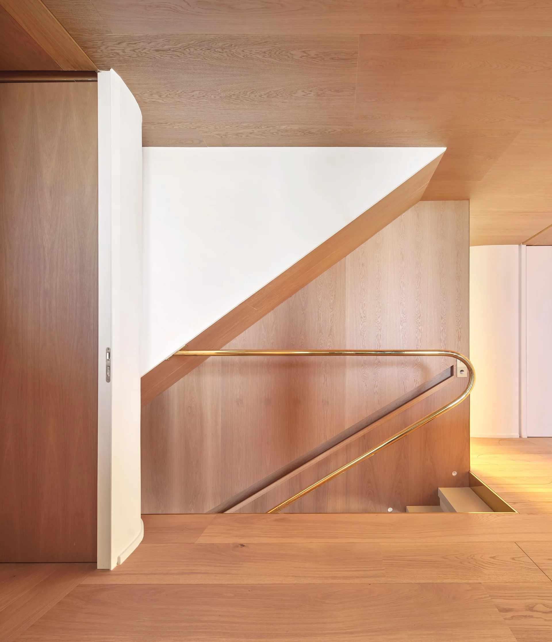 A modern staircase lined with warm oak wood paneling on the walls and ceiling, featuring a polished curved brass handrail.